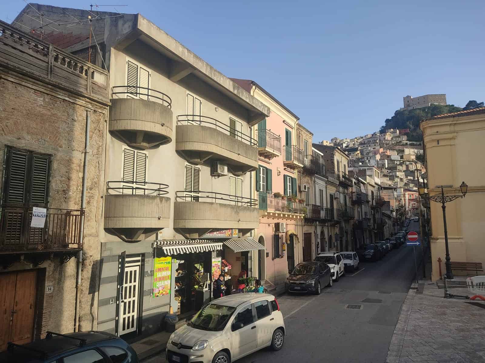 Vani di immobili in stile mediterraneo, con edifici storici e moderni, strada affascinante in centro città, vista sul castello sulla collina, appartamenti con balconi e negozi di vendita e affitto, atmosfera autentica.