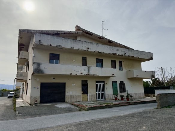 Una casa fronte strada con problemi di degrado e danni alla facciata, in un contesto urbano con poche piante, pronta per ristrutturazione o ricostruzione nel settore immobiliare NextCasa.