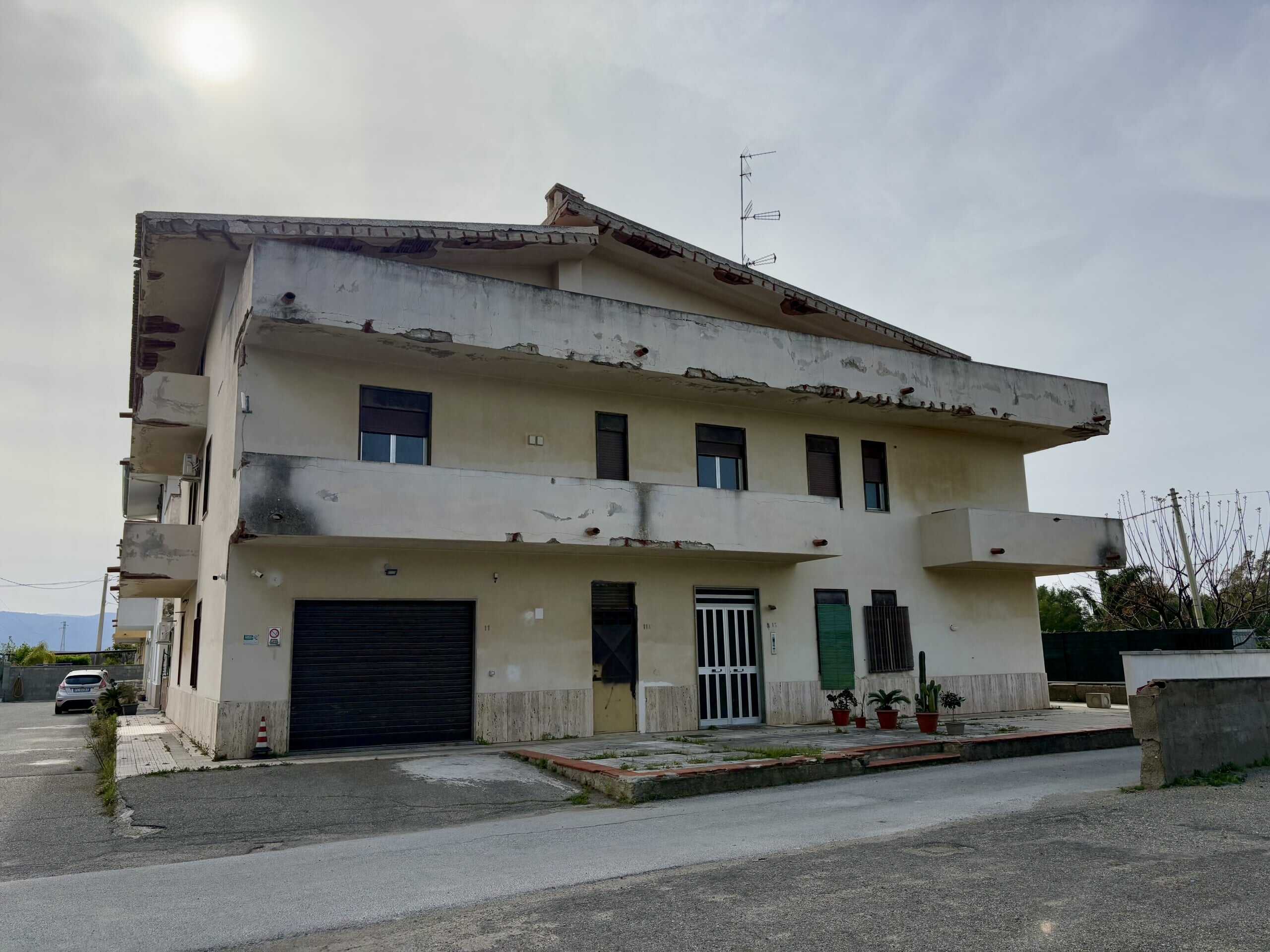 Una casa fronte strada con problemi di degrado e danni alla facciata, in un contesto urbano con poche piante, pronta per ristrutturazione o ricostruzione nel settore immobiliare NextCasa.