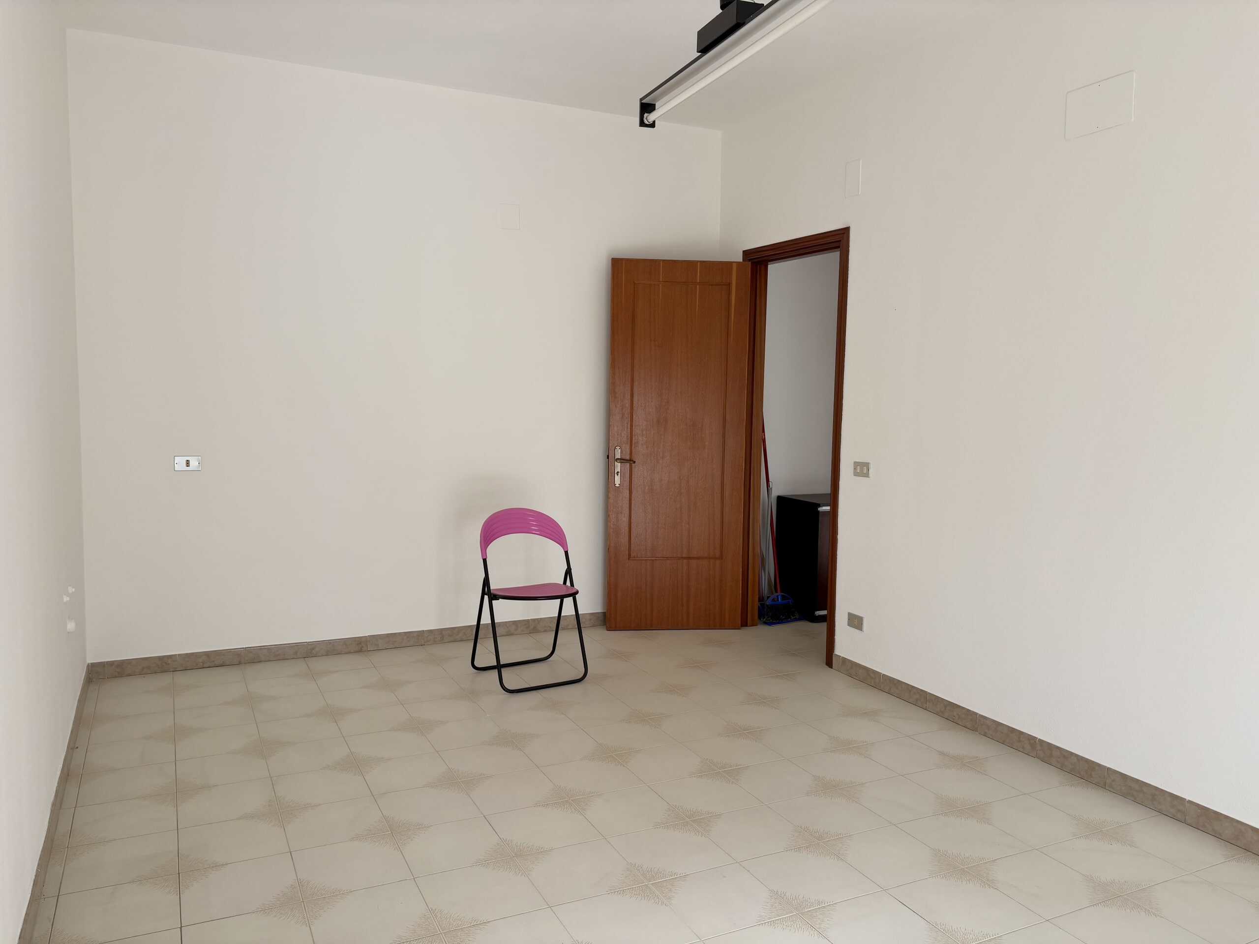 Sedie rosa in stanza vuota con pareti bianche e pavimento in piastrelle beige con motivo, porta di legno aperta, plafoniera a binario, spazio per arredi e real estate a Napoli.