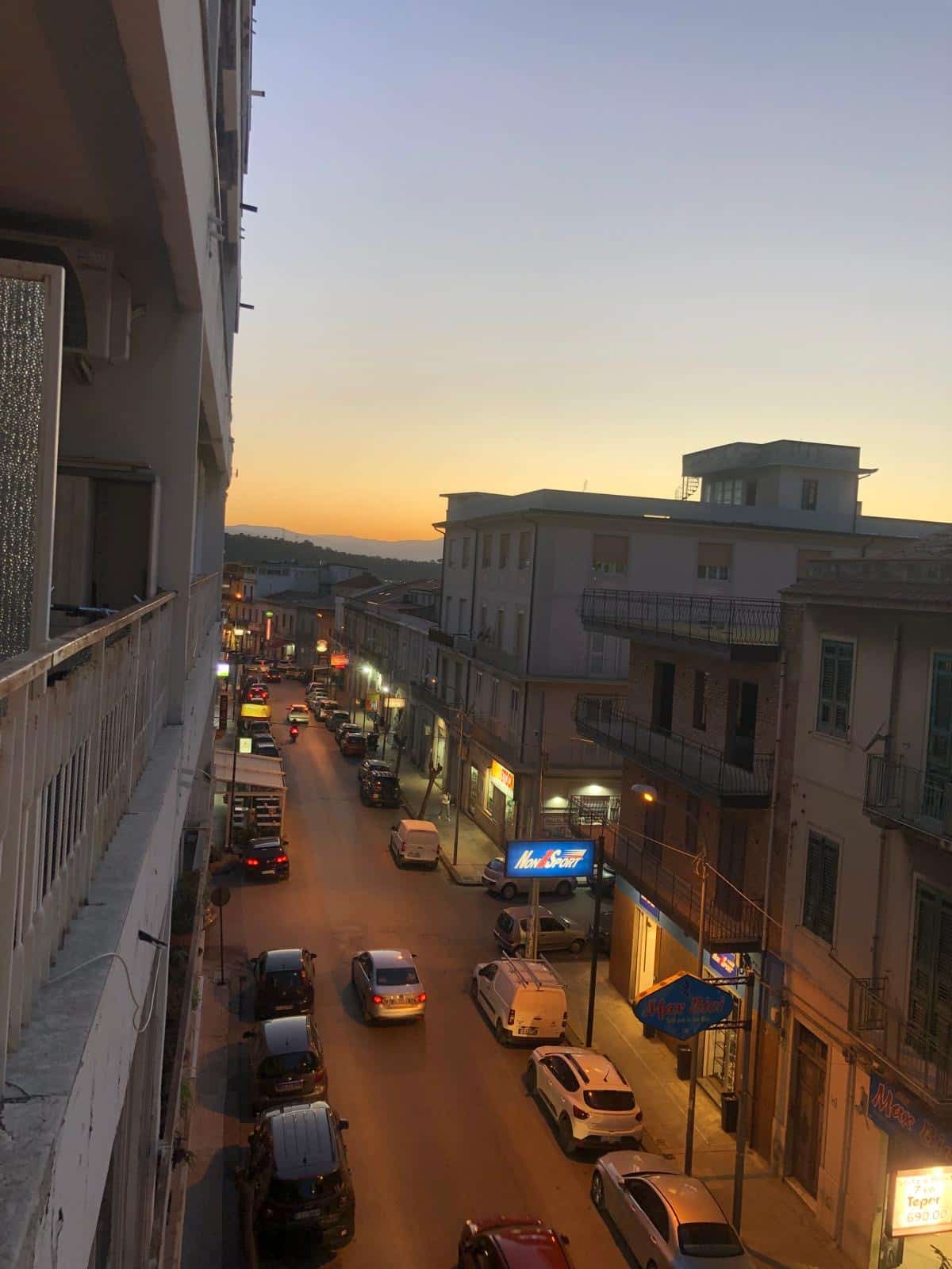 Veduta urbana al tramonto con strada principale, negozi e auto parcheggiate, atmosfera vivace e tranquilla, illuminazione serale, edificio storico e cielo al crepuscolo.