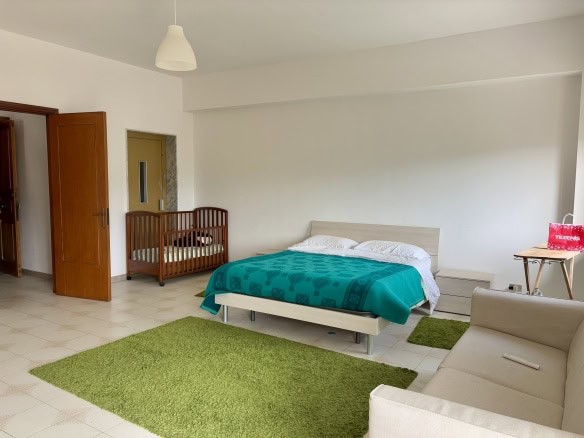 Spazio soggiorno con letto matrimoniale, culla per neonati e zona relax in appartamento NextCasa, stile minimalista, arredato con mobili moderni, tappeti verdi e ambienti luminosi.
