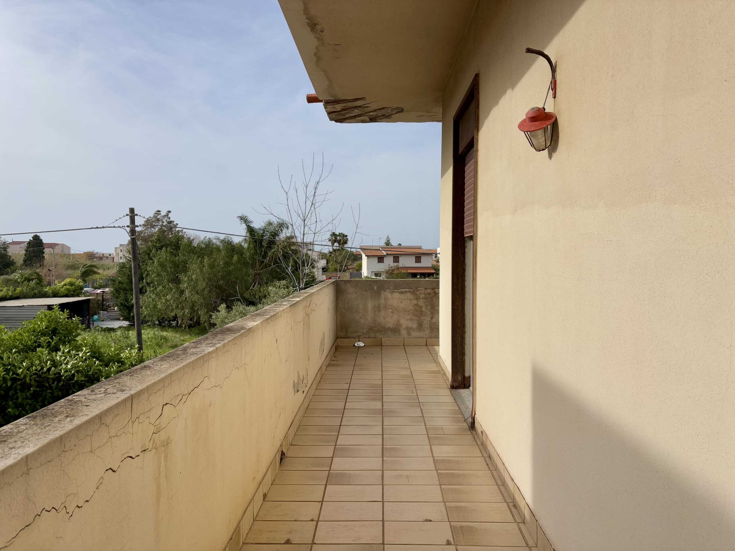 Balcone spazioso e luminoso di un appartamento, con vista su alberi e case, pareti beige, griglie di protezione, luci esterne e pavimento in piastrelle, ideale per relax e momenti all'aperto.