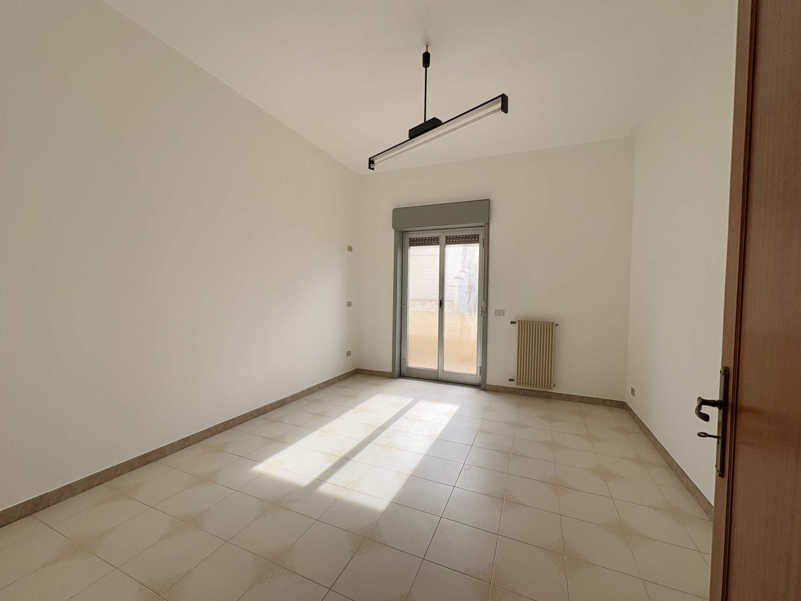 Ampio stanza vuota con parquet chiaro, grande finestra con accesso al balcone, pareti bianche, riscaldamento a radiatore, illuminazione moderna a soffitto, ideale per appartamenti in vendita o affitto, ambiente luminoso e accogliente.