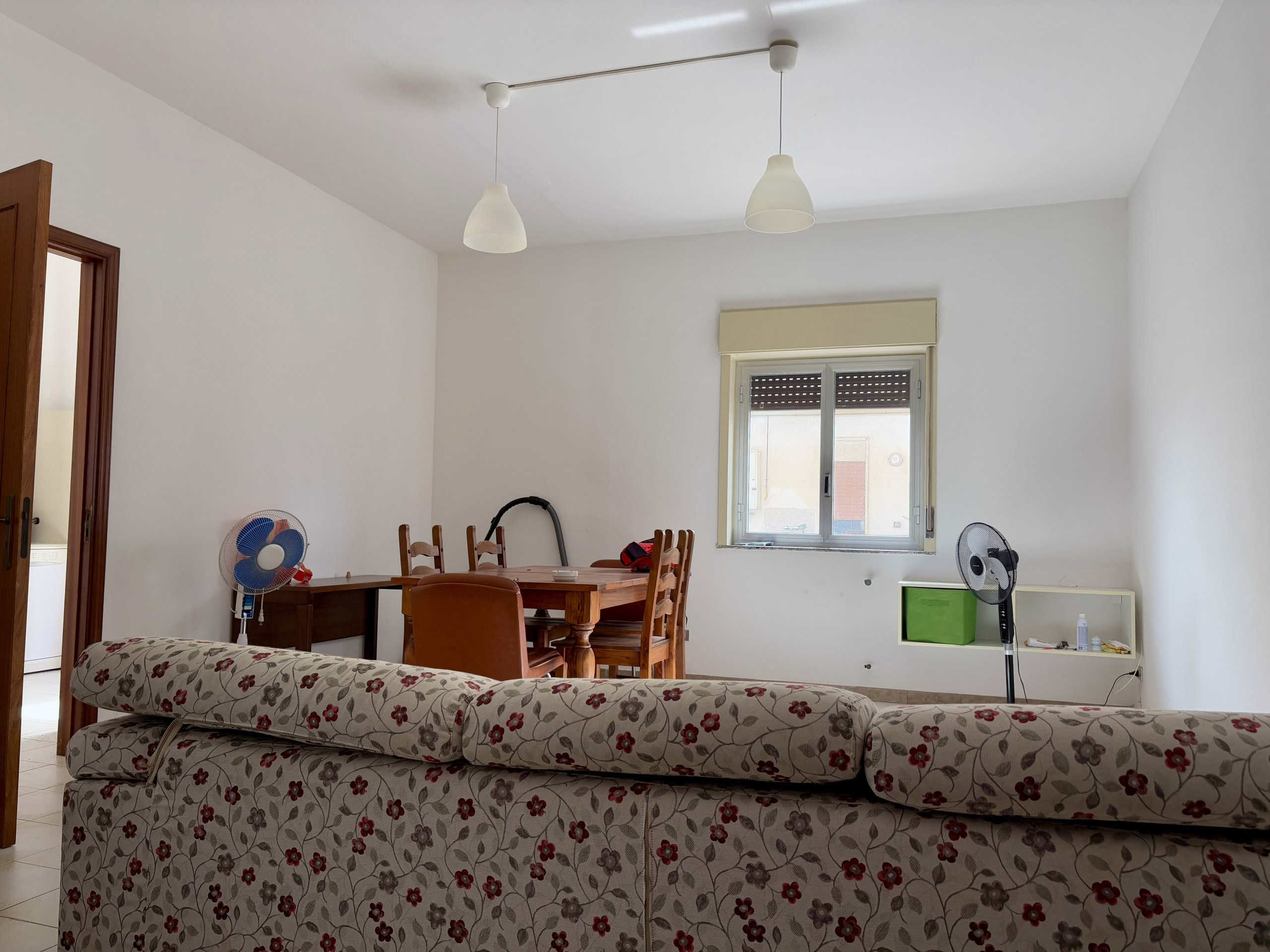 Fantasia soggiorno con divano floreale, tavolo da pranzo con sedie in legno, finestra con tapparella, ventilatori portatili, ambiente luminoso e spazioso.