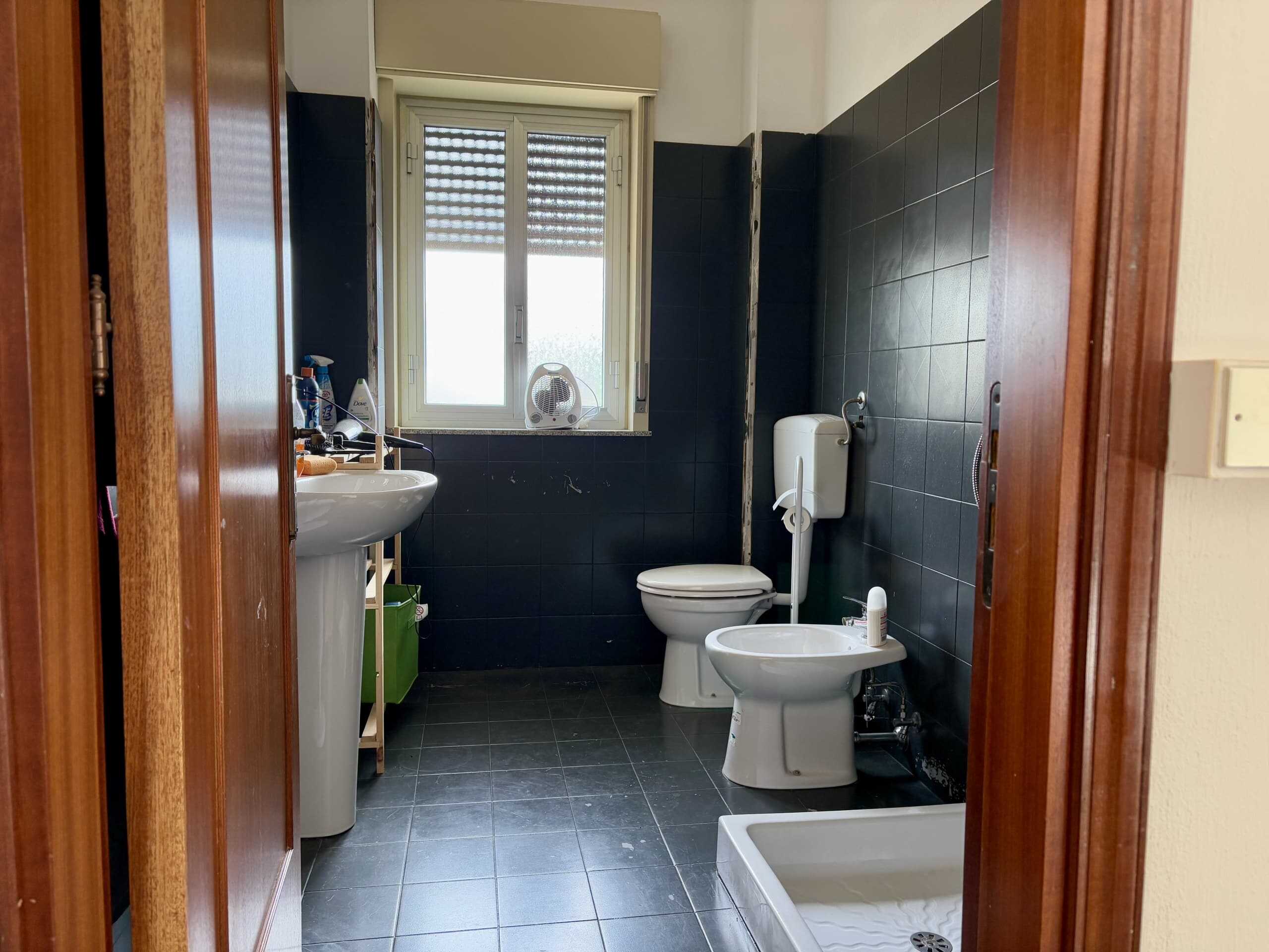 Lavabo, bidet e wc in bagno di casa con finestra, piastrelle nere e arredi di legno, vista dall'ingresso, in stile semplice e funzionale, in zona residenziale.