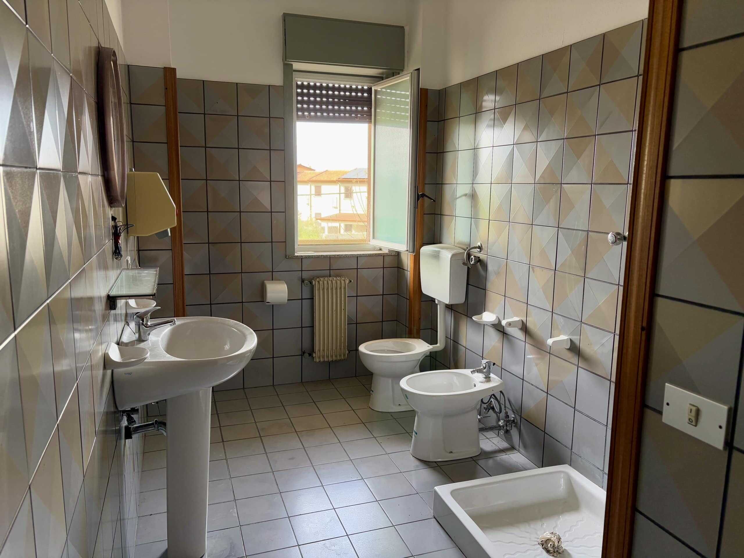 Lavabo, bidet, water closet, e doccia in bagno spazioso con pareti rivestite di piastrelle beige e grigie, finestra aperta che permette l'ingresso di luce naturale, ideale per soluzioni immobiliari di NextCasa.
