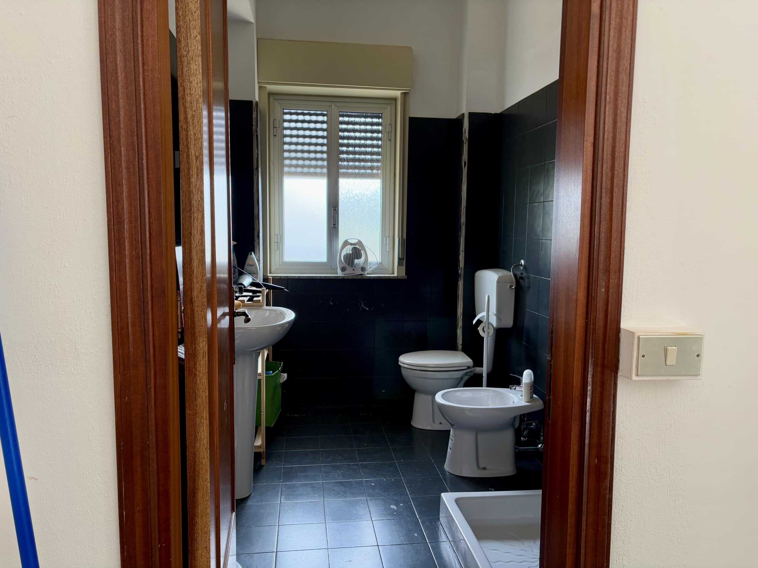 Lavabo e gabinetto in bagno di appartamento con finestra, piastrelle nere, sanitari bianchi e bidet, vista dall'ingresso, stile semplice e funzionale, ambiente luminoso e pratico.