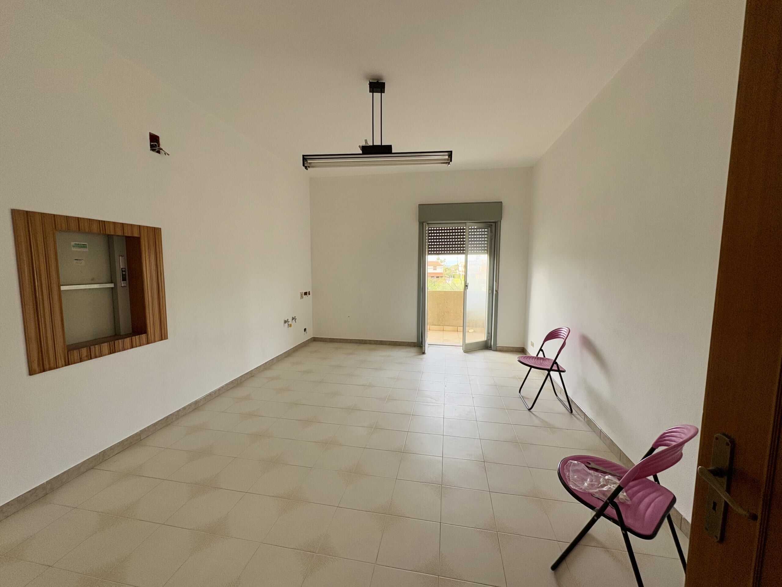 Soggiorno vuoto con porte finestra su balcone, pareti bianche, piastrelle beige, due sedie pieghevoli rosa e soffitto con lampadario lineare.