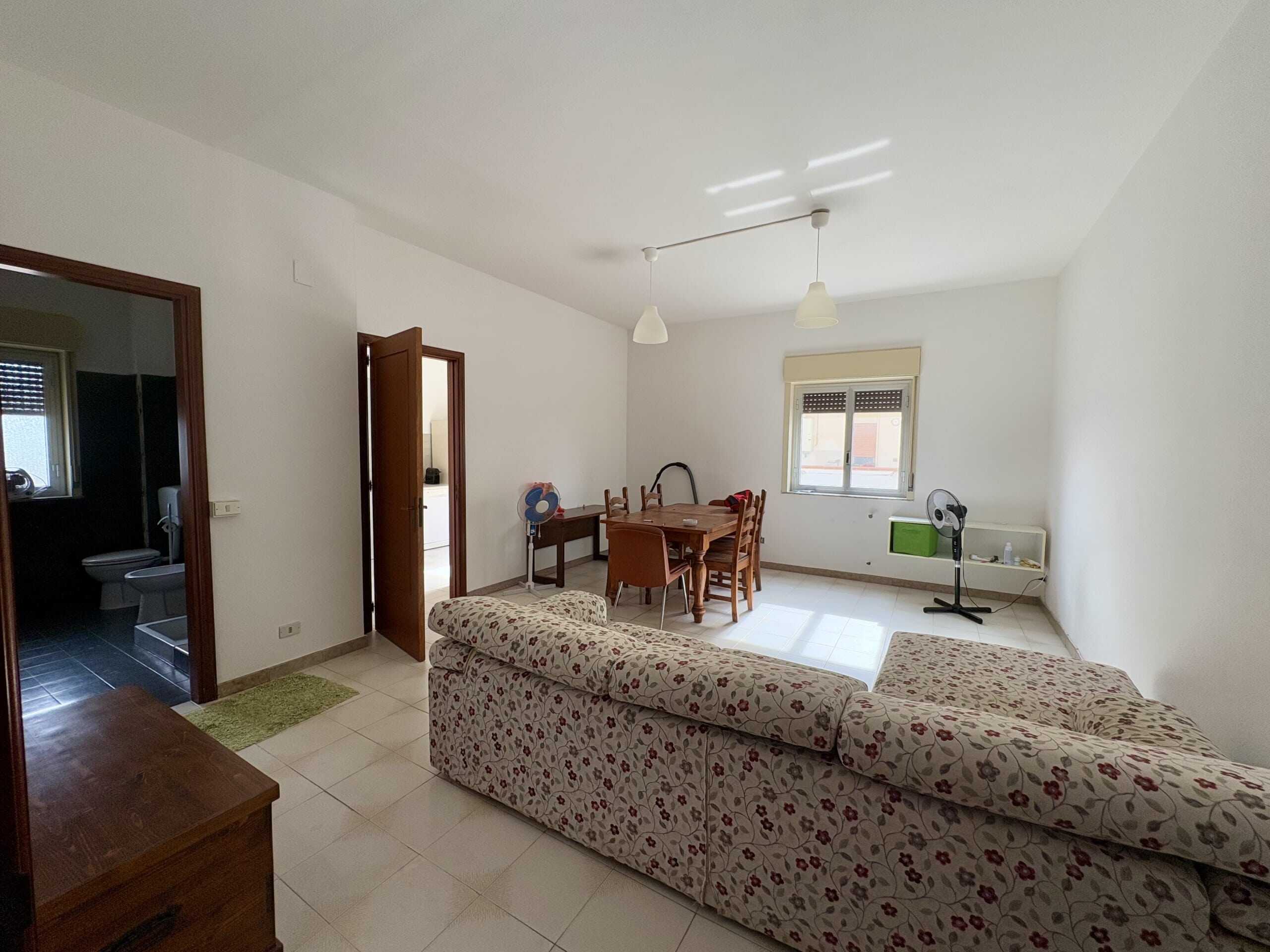 Soggiorno spazioso con divano a fiori, tavolo da pranzo in legno, finestre luminose, aria condizionata e ventilatori, ideale per vivere in casa con comfort e luminosità.