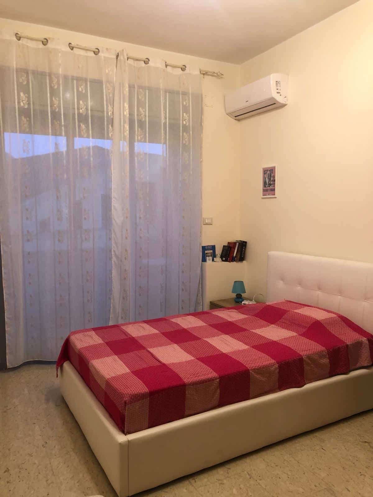 Elegante camera da letto con letto moderno, copertura a quadri rosso e rosa, finestra con tende floral e atmosfera accogliente, ideale per immobili in vendita o affitto da NextCasa.