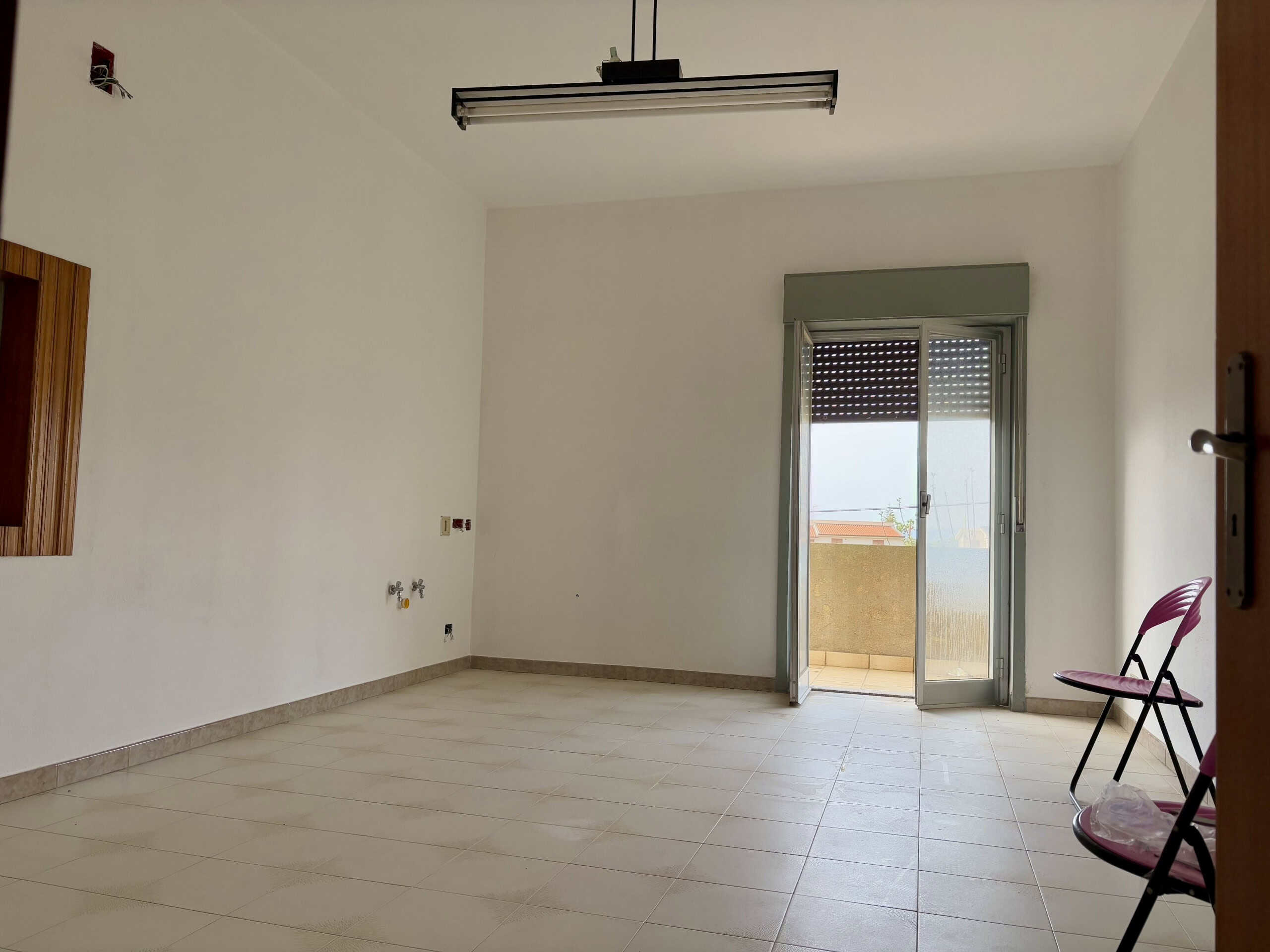 Ampia stanza vuota con porta finestra che dà su un balcone, pareti bianche e pavimento in piastrelle beige, ideale per l'arredamento di case in vendita o affitto NextCasa.