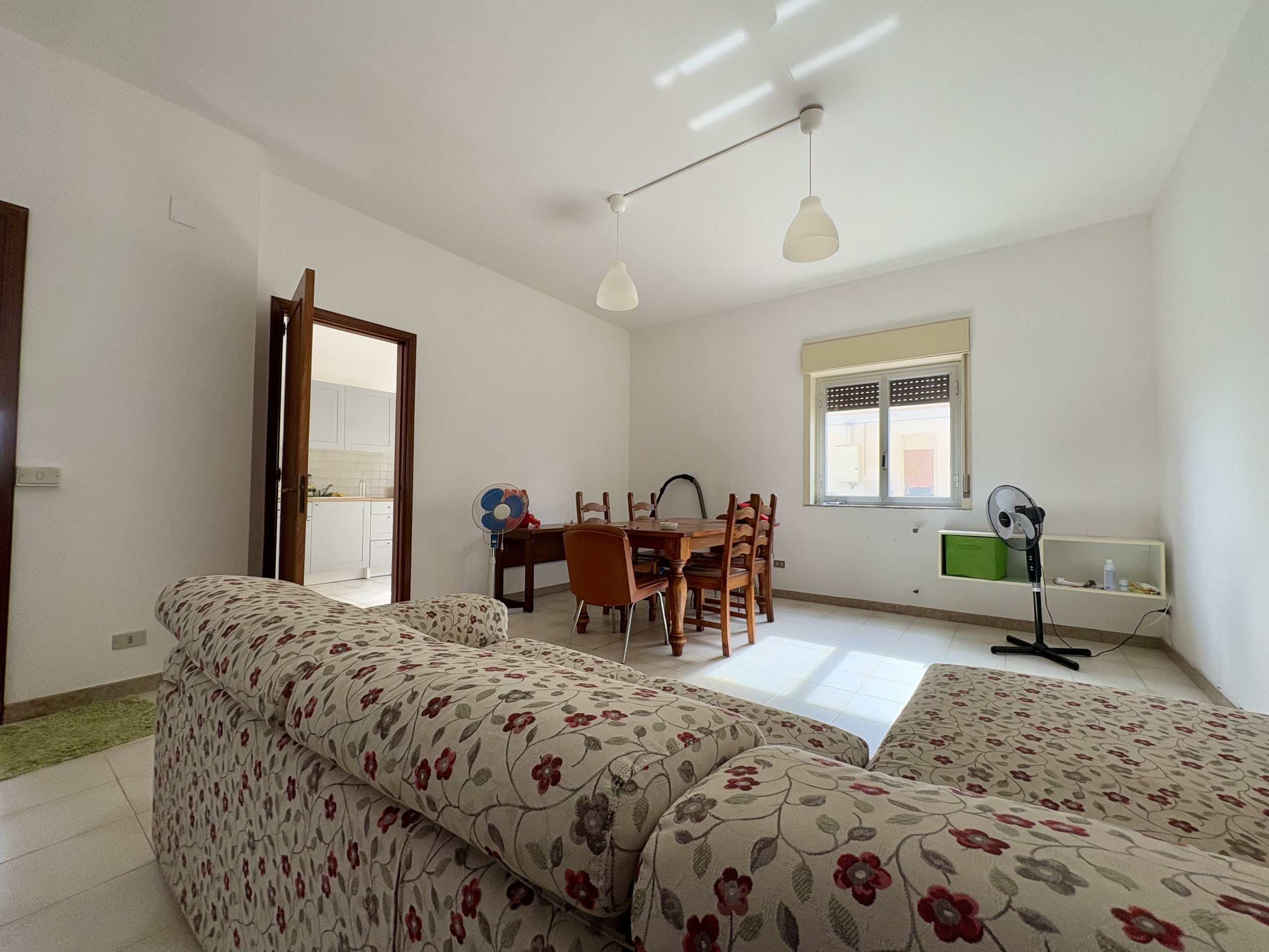 Spazio soggiorno con divano fiorato, tavolo da pranzo e accessori, interni luminosi, finestra con tende, ambiente abitabile e confortevole.