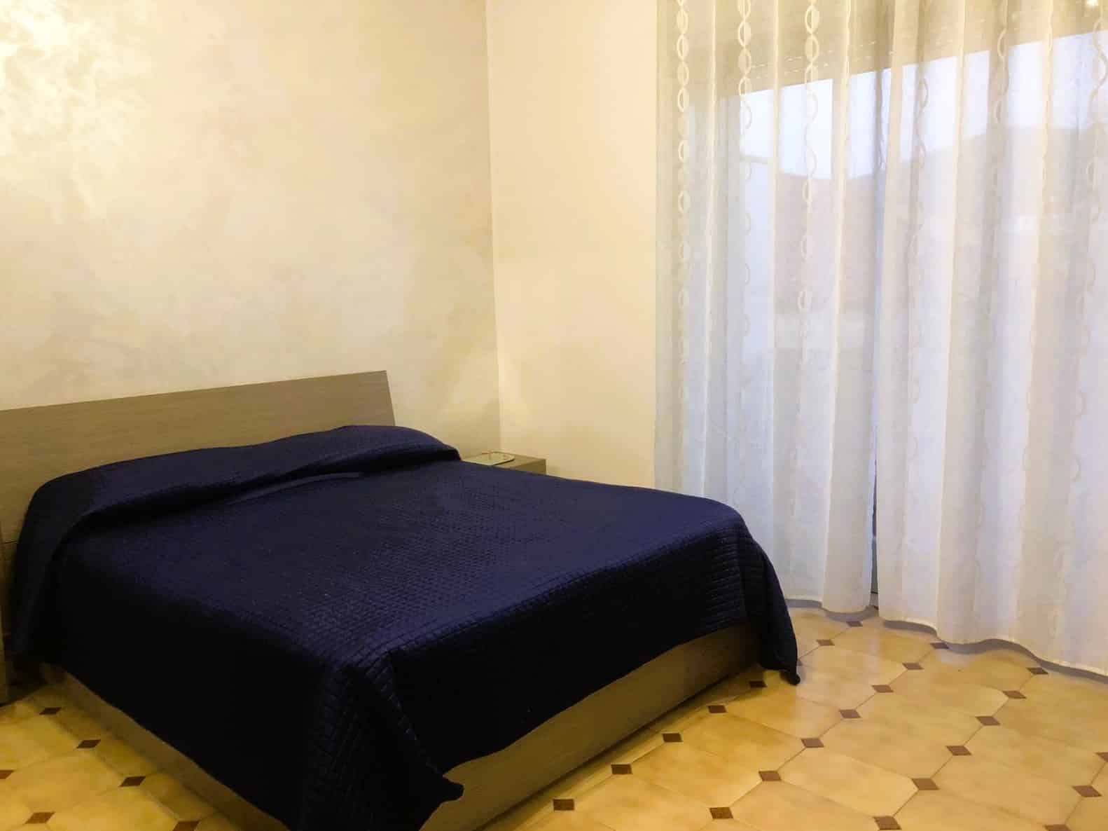 Letto matrimoniale in camera elegante con tende di fronte a finestra luminosa, arredamento moderno, pavimento in legno chiaro, atmosfera accogliente per soggiorni confortevoli.