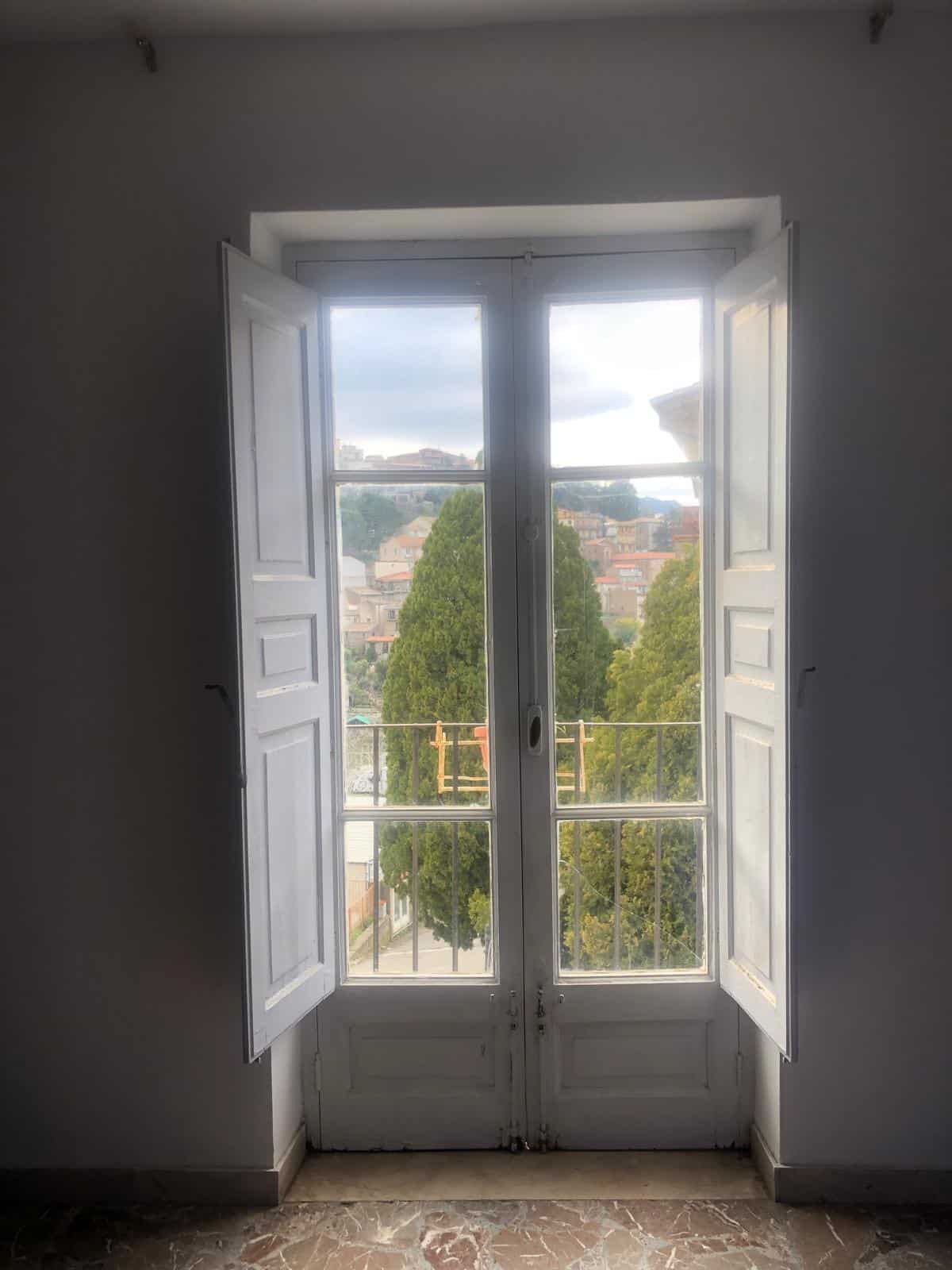 Vista di una finestra con persiane bianche che si apre su un panorama collinare con case e alberi, illuminando l'interno di una stanza in stile classico.