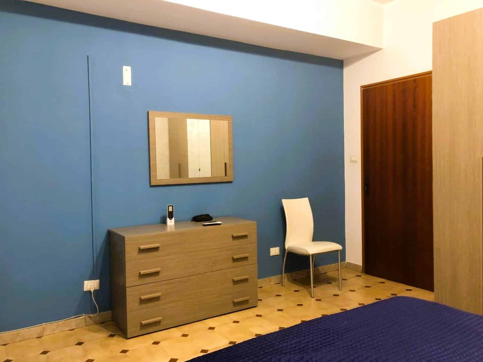 Blue wall bedroom interior with wooden dresser, mirror, white chair, and parquet flooring in modern apartment.