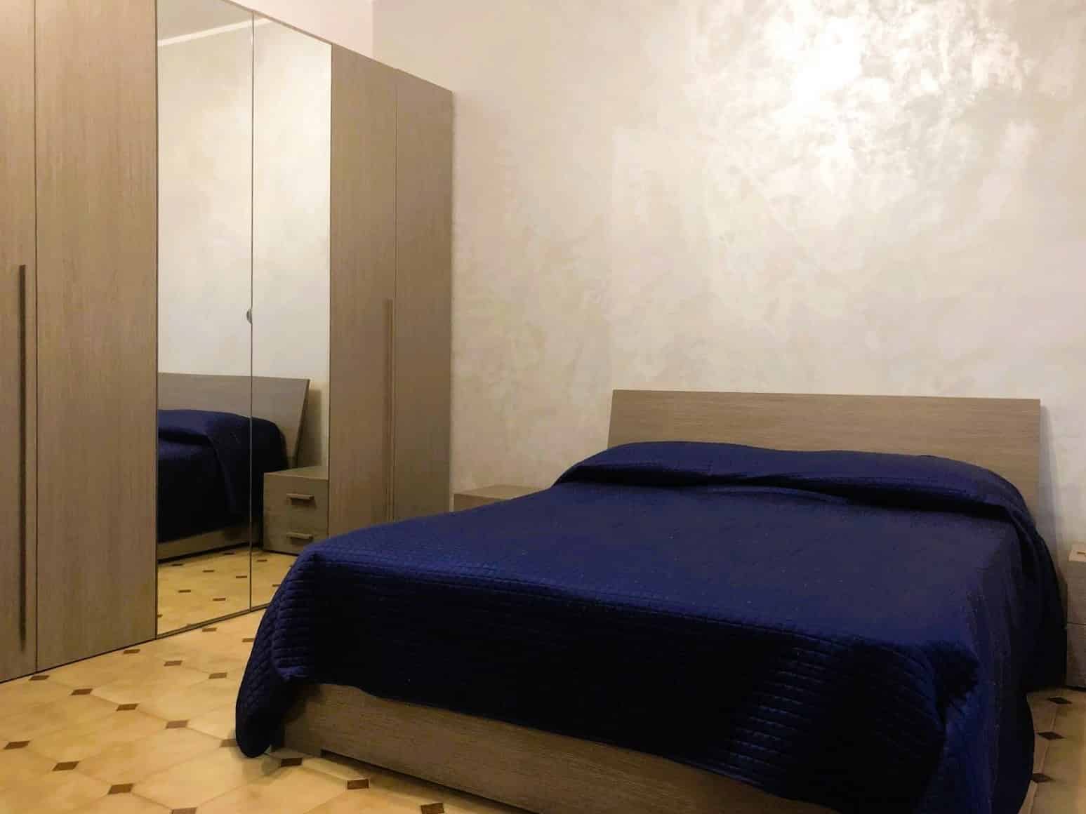 Letta camera da letto con letto matrimoniale, lenzuola blu, armadio con ante a specchio, pareti beige e pavimento in legno chiaro, ambiente accogliente e moderno.
