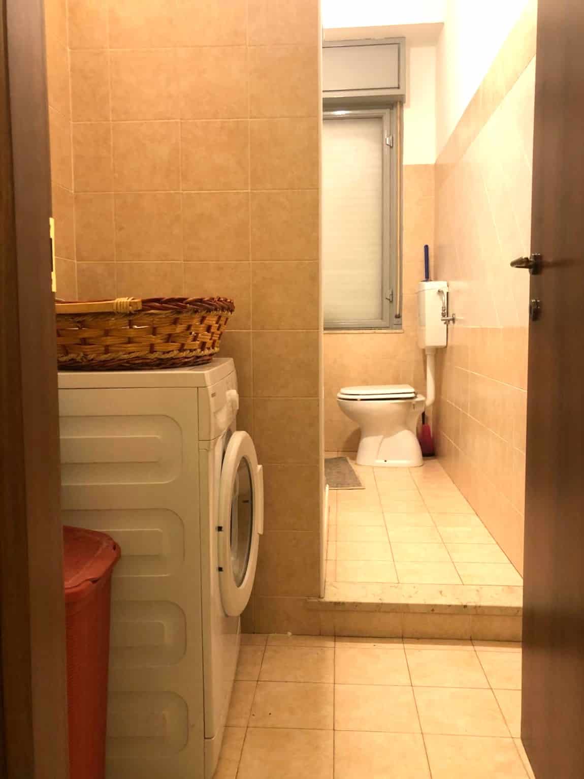 Lavatrice con basket in ingresso bagno con sanitari e finestra, rivestimenti in ceramica beige, ambiente funzionale e pratico, ideale per appartamenti e case.