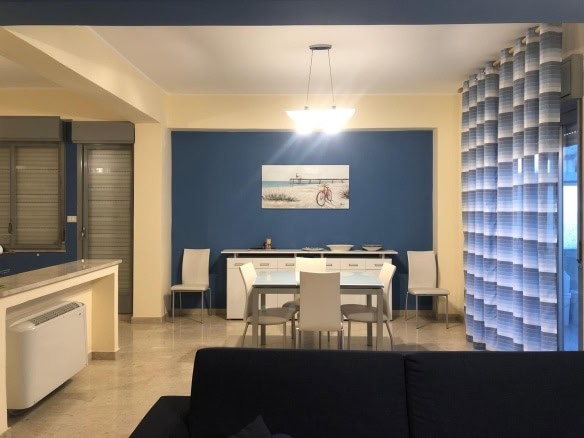 Luminoso soggiorno con parete blu, tavolo bianco e sedie moderne, decorazioni marine, grandi finestre con tende a strisce blu e bianche, illuminazione centrale, ambiente accogliente e luminoso.