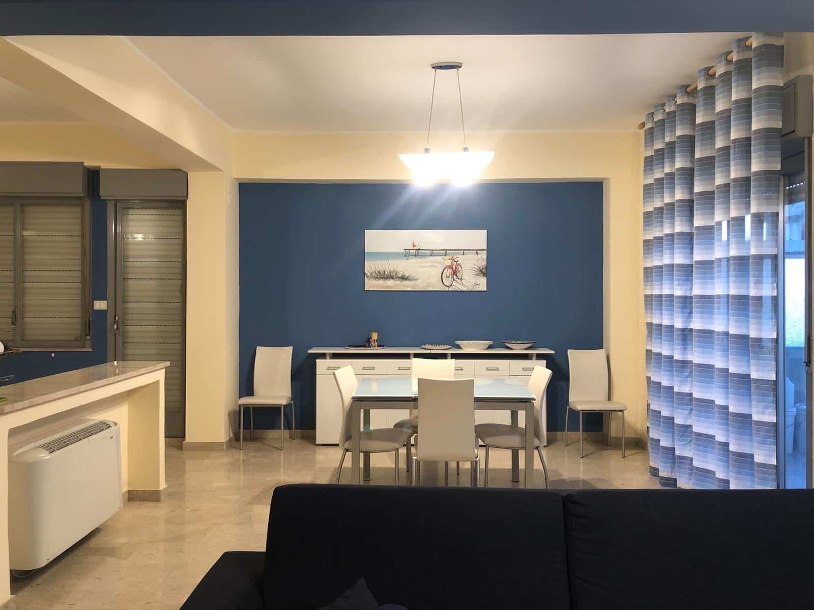 Luminoso soggiorno con parete blu, tavolo bianco e sedie moderne, decorazioni marine, grandi finestre con tende a strisce blu e bianche, illuminazione centrale, ambiente accogliente e luminoso.