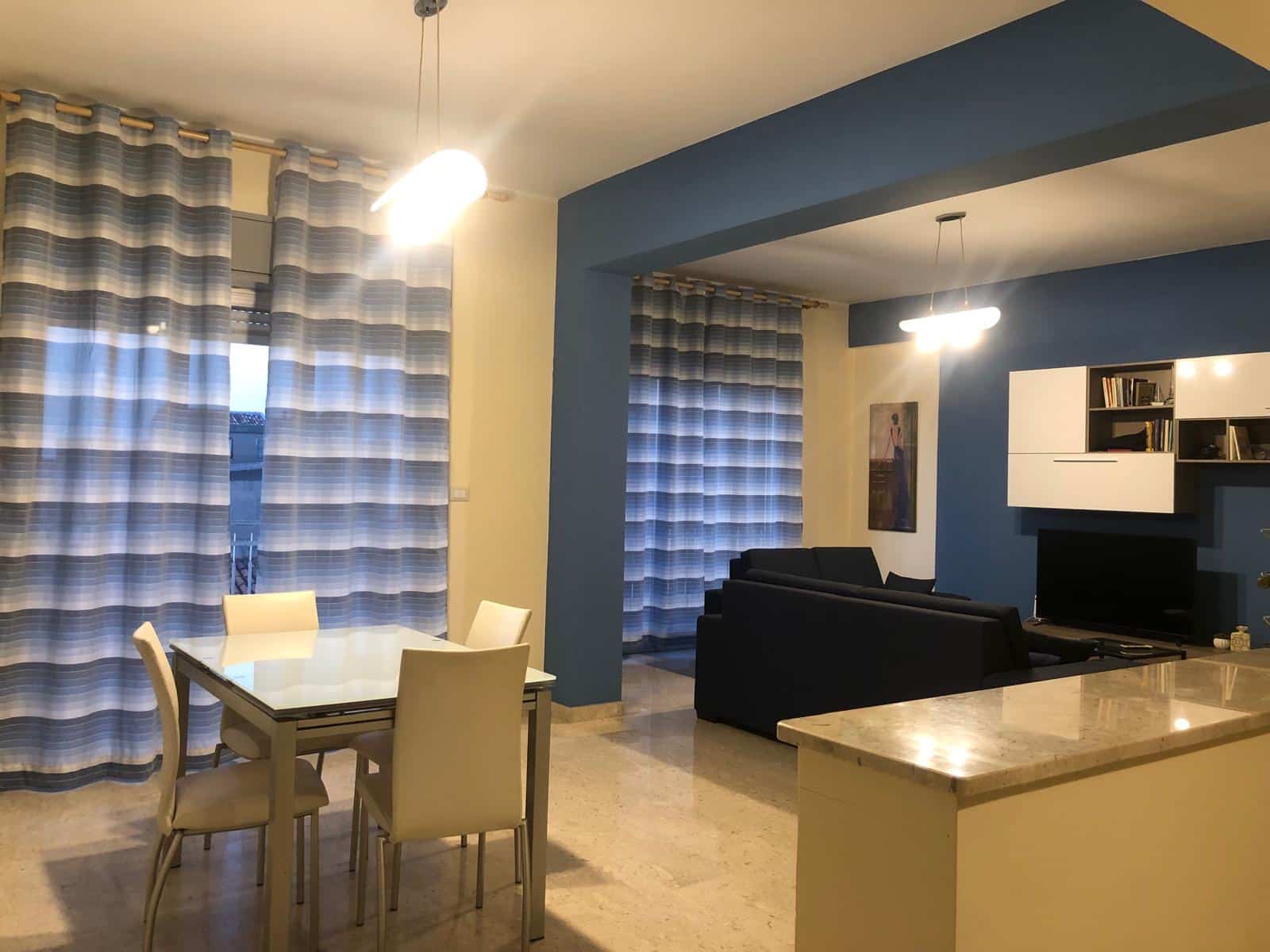 Elegante soggiorno con zona pranzo e area relax, arredato in stile moderno, pareti blu e illuminazione soffusa, ideale per vivere momenti di comfort e convivialità con vista luminosa su ampie vetrate con tende a righe.