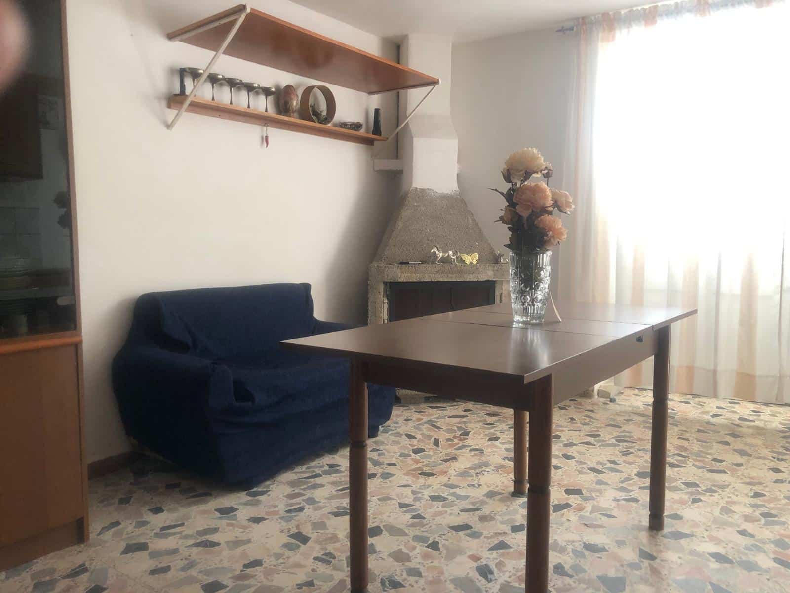 Soggiorno con camino in pietra, divano blu, tavolo in legno con vaso di fiori freschi, pareti chiare e tendaggi leggeri, ambiente accogliente e luminoso, ideale per case in vendita o affitto NextCasa.