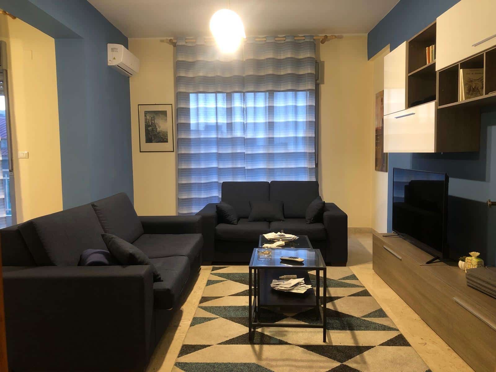 Soggiorno moderno con divani neri, televisore a schermo piatto e mensole arredate, ambiente accogliente e luminoso con tende a righe e pareti colorate blu e giallo.