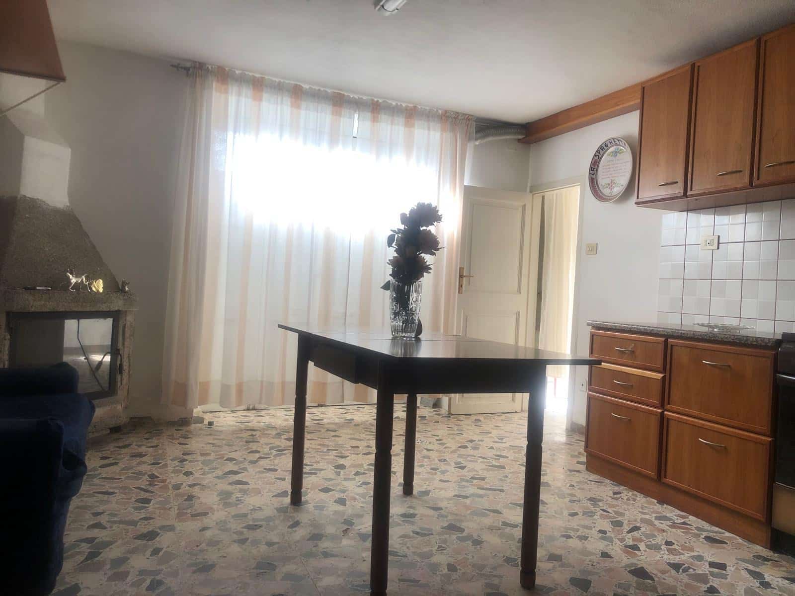 Finestra luminosa in cucina con tende leggere, arredamento in legno, tavolo al centro con vaso di fiori, camino in pietra, pavimento in marmo, interni in stile classico, ambiente caldo e accogliente.
