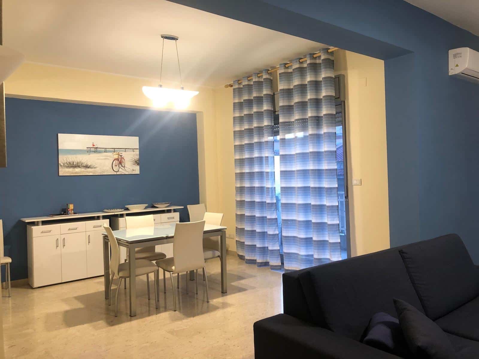 Accogliente soggiorno con parete blu, grande finestra con tende a righe e zona pranzo moderna. Ambiente luminoso e confortevole, ideale per famiglie o coppie, perfetto per vivere momenti di relax e convivialità.
