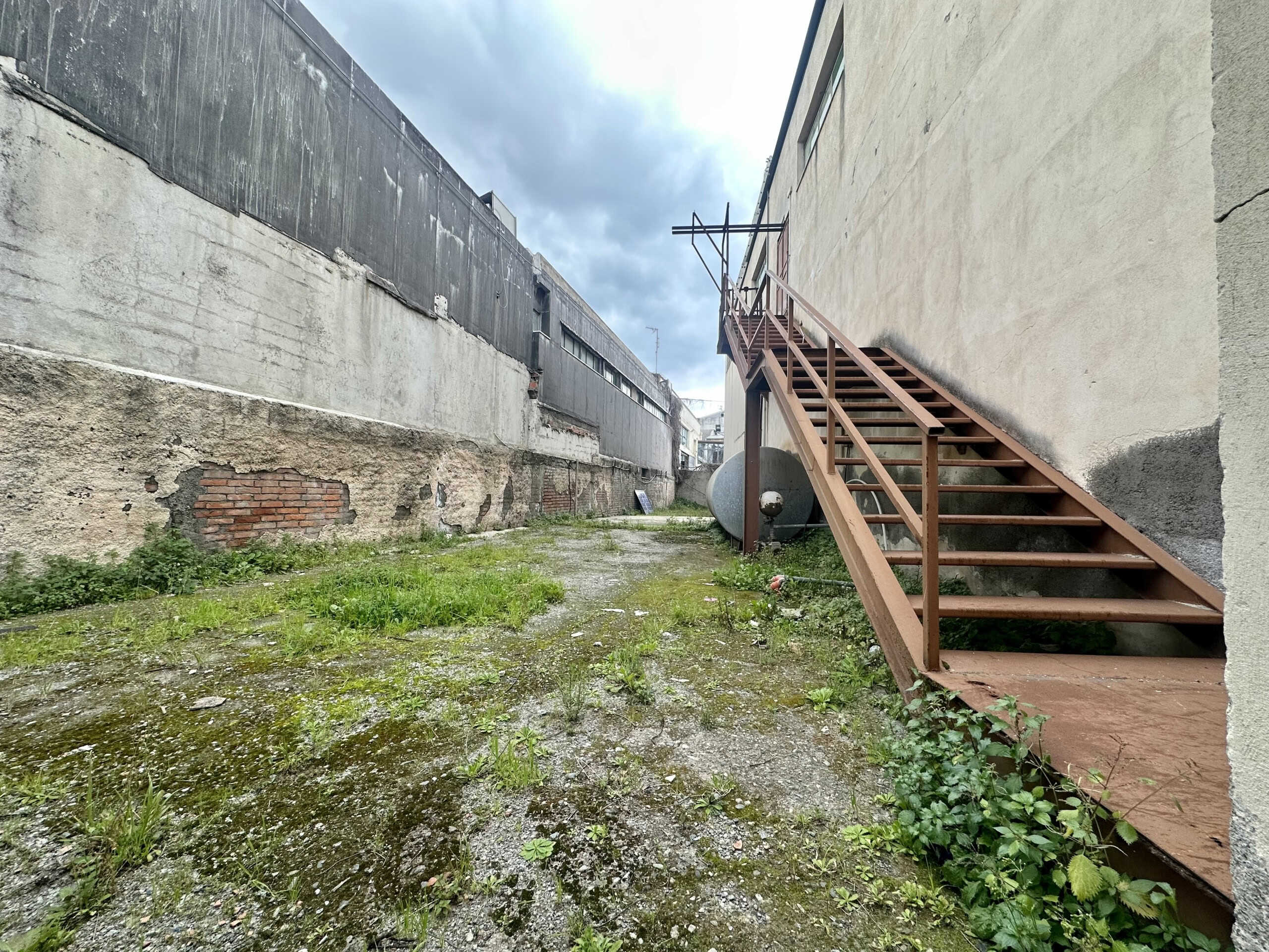 Scalinata esterna in ferro in un cortile in pietra con pareti di cinta in condizioni di abbandono, situata in un contesto urbano.