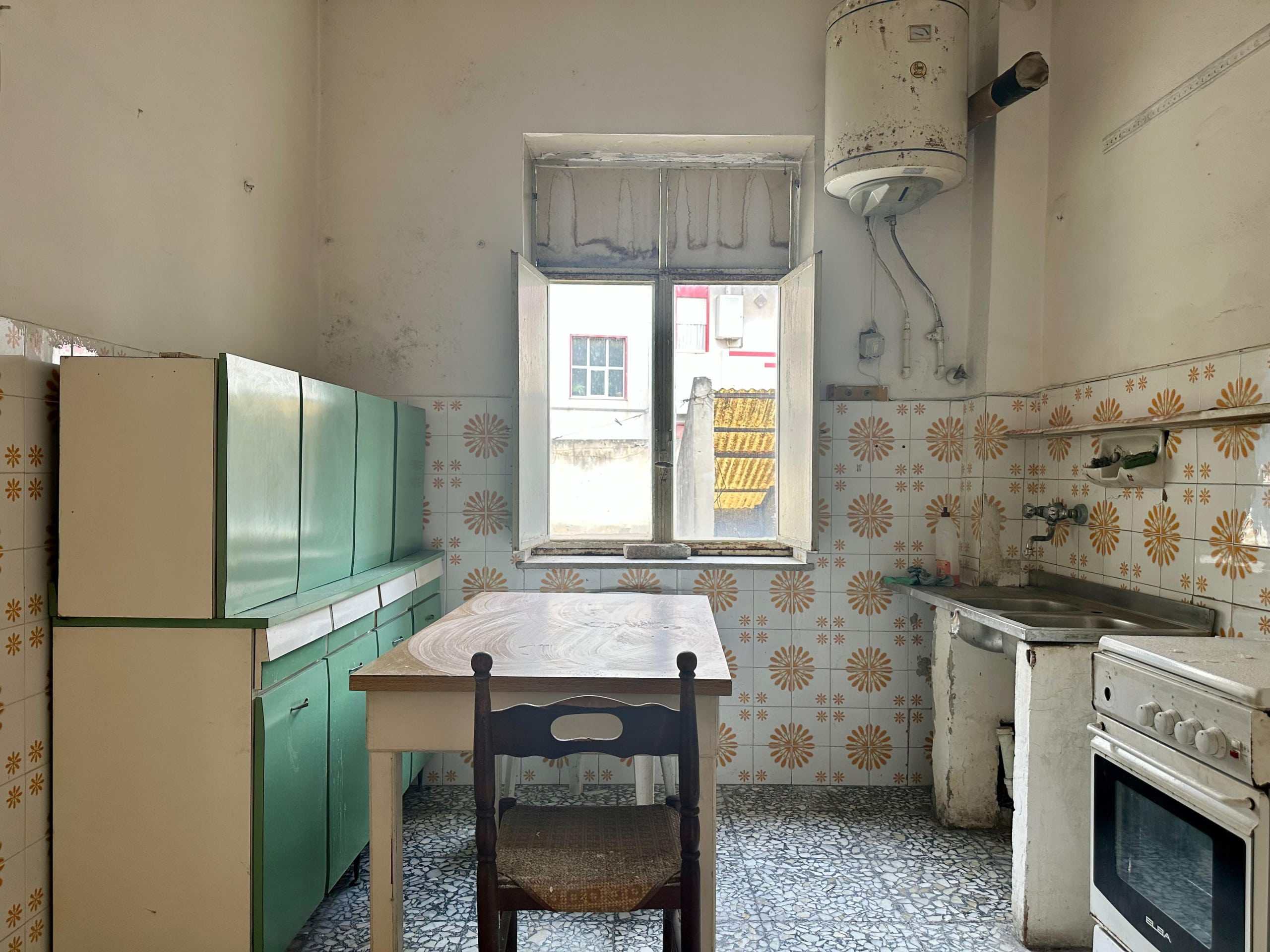 Arredo cucina antico, cucina in stile vintage, stanza cucina ristrutturata, aria luminosa, ambiente rustico e funzionale, spazio abitativo da rinnovare, dettagli di piastrelle floreali, comfort semplice e accogliente, cucina con forno e lavabo, vista sulla città dall'apertura della finestra.