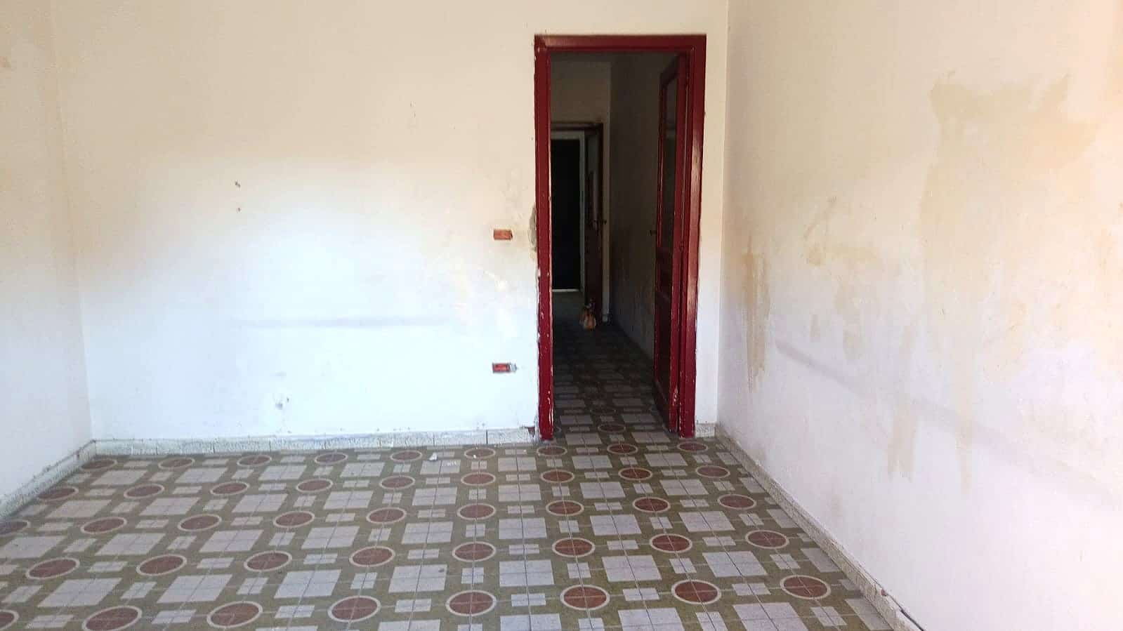Vecchia stanza da ristrutturare con pareti bianche e piastrelle decorative sul pavimento, ingresso a soffitto alto e porta rossa, pronta per essere ristrutturata e personalizzata.