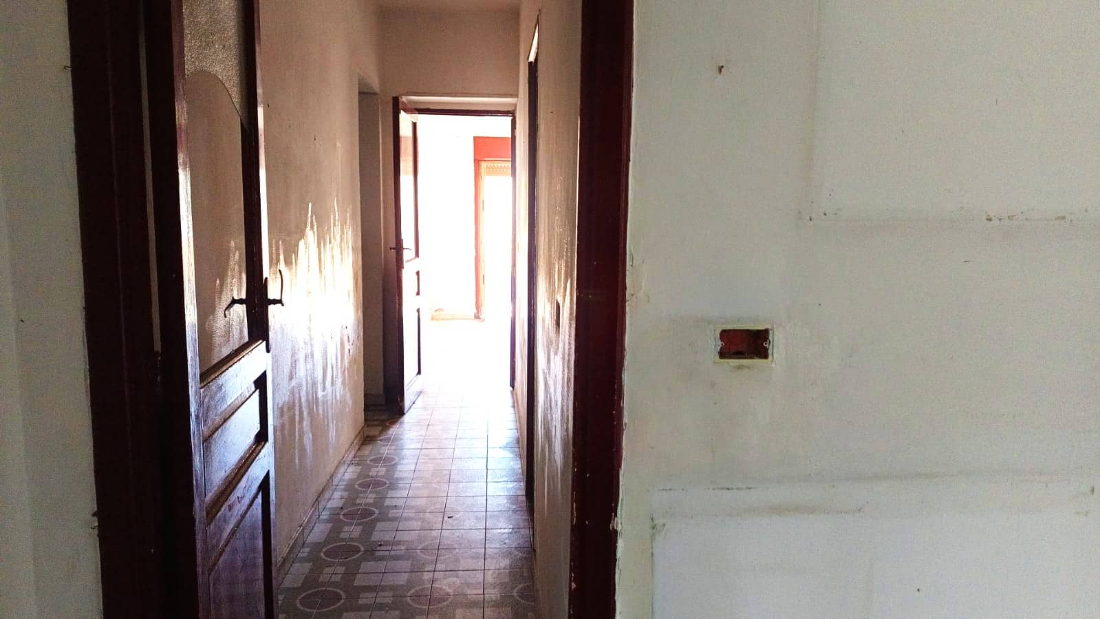 Porta di legno in stile classico, corridoio interno di casa da rinnovare, pavimentazione in piastrelle geometriche, pareti bianche con segni di usura, ambiente abitativo da ristrutturare, immagine di un immobile in vendita o affitto, NextCasa, agente immobiliare specializzato in ristrutturazioni e compravendite immobiliari.