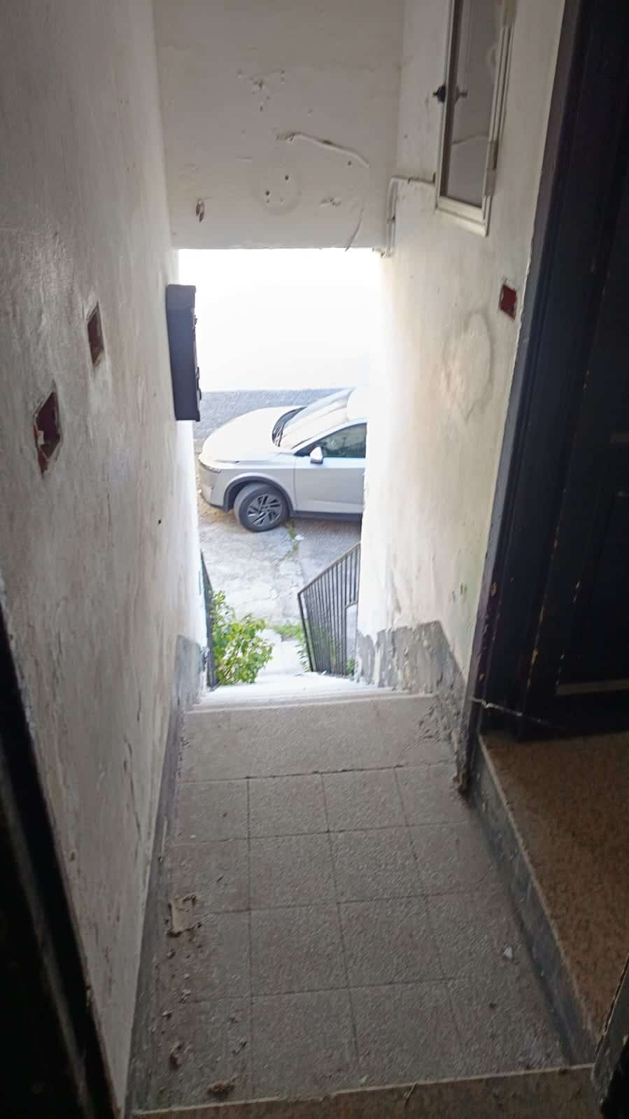 Portico con scala esterna e vista sulla strada con auto parcheggiate, ingresso da ristrutturare, condizioni di abbandono, lapidi grigie e pareti con deterioramento, ambiente da rinnovare, immobile da sistemare, spazio di accesso esterno.