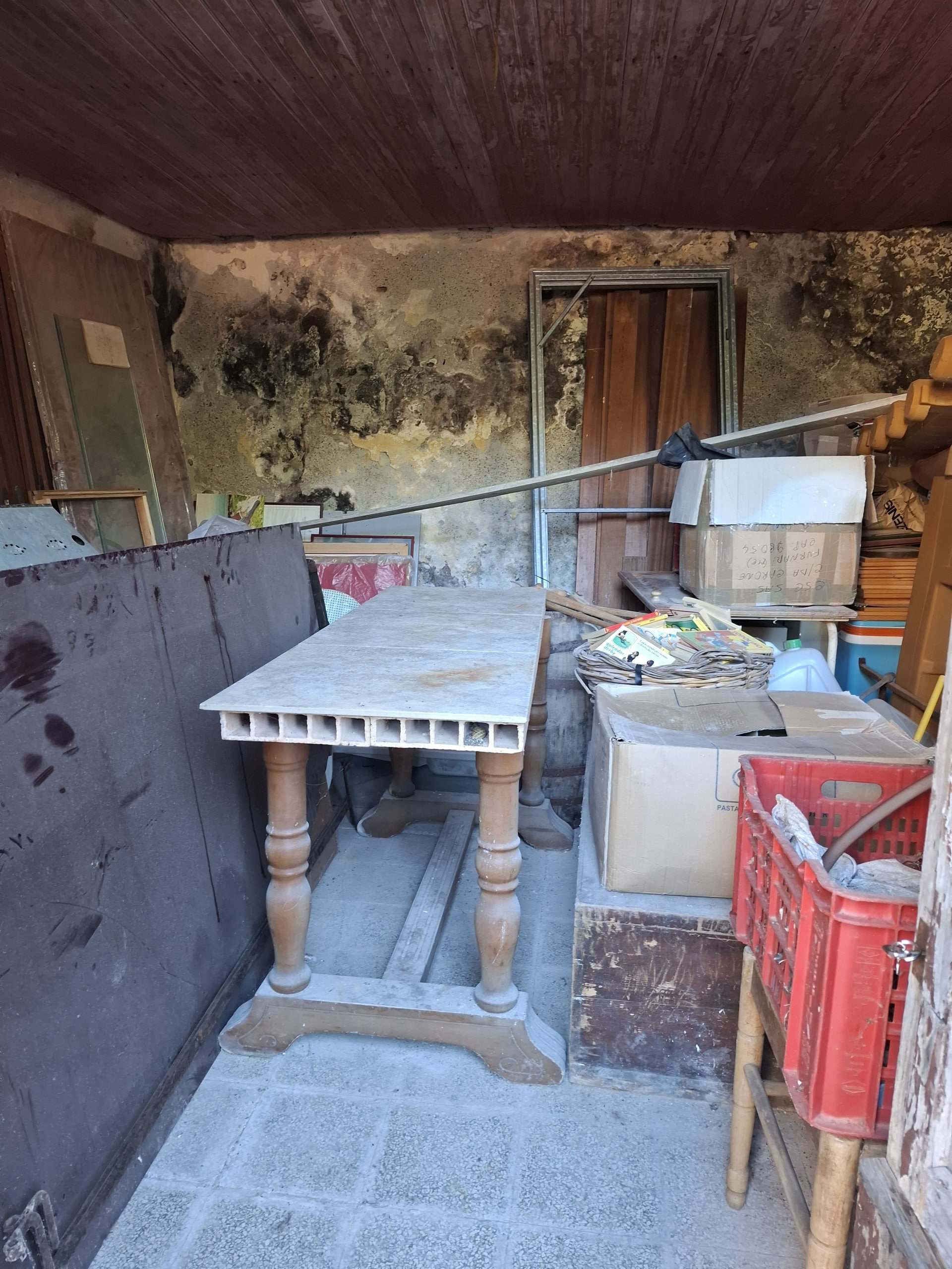 Vecchio tavolo in legno con gambe tornite, spazio di stoccaggio sovraccarico e pareti con muffa e umidità, ambiente di deposito o cantina con oggetti vari.