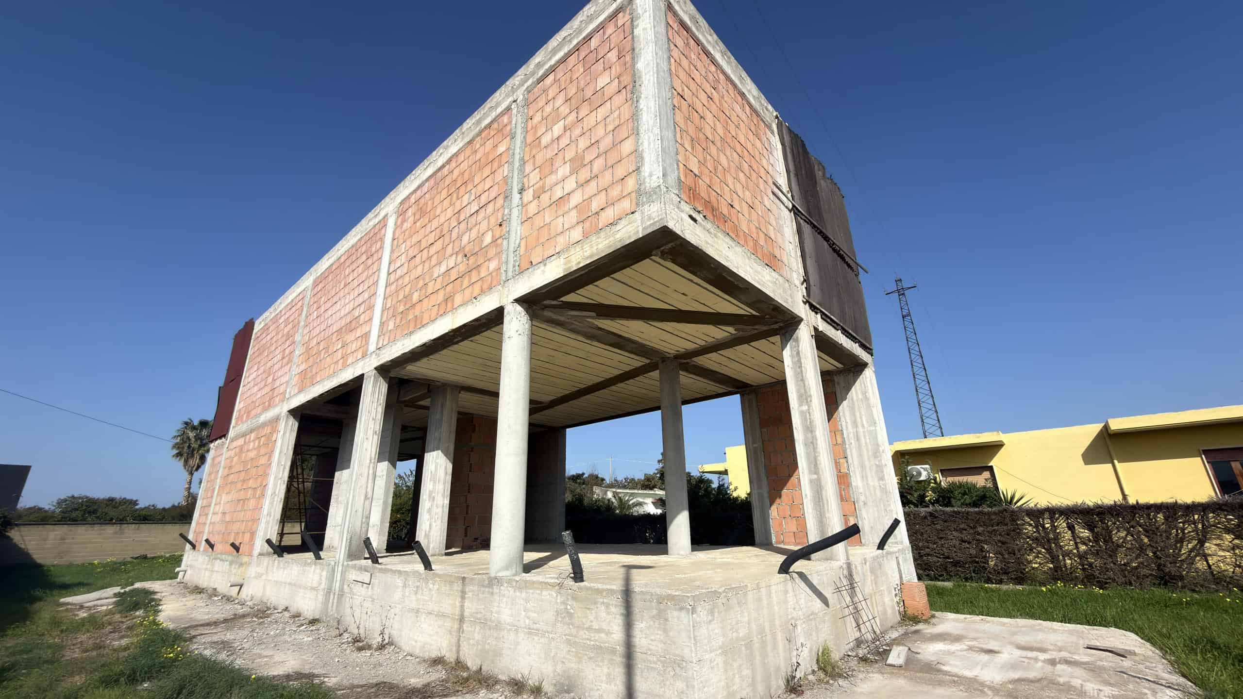 Pannello di costruzione di una casa in fase di realizzazione con struttura in cemento e mattoni, in un'area aperta soleggiata con cielo blu.