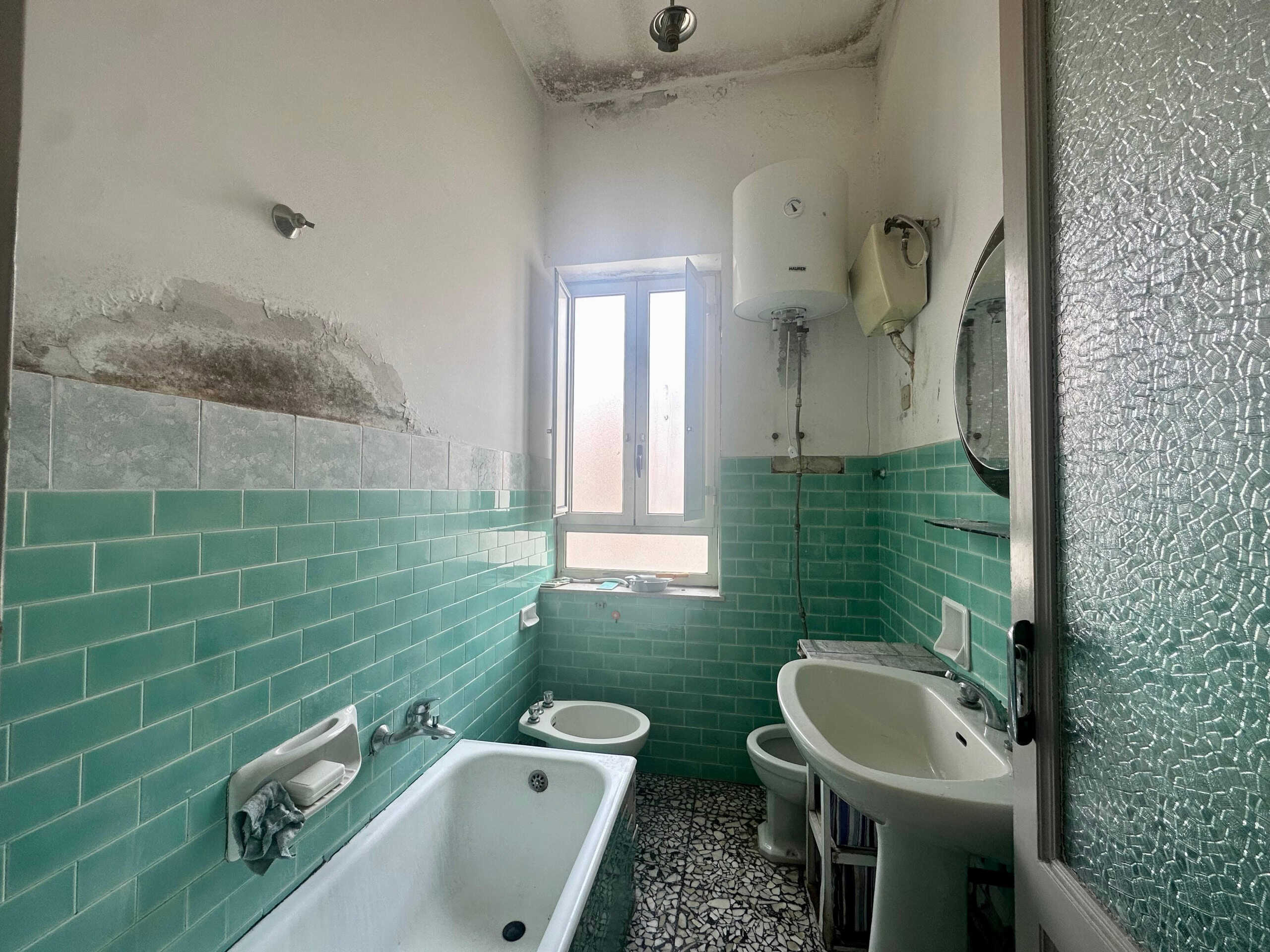 Bagno in fase di ristrutturazione con piastrelle verdi, vecchio pavimento in terrazzo, servizi igienici e punto acqua, finestra per luce naturale, pareti da rinnovare, ambiente abitabile per una ristrutturazione.