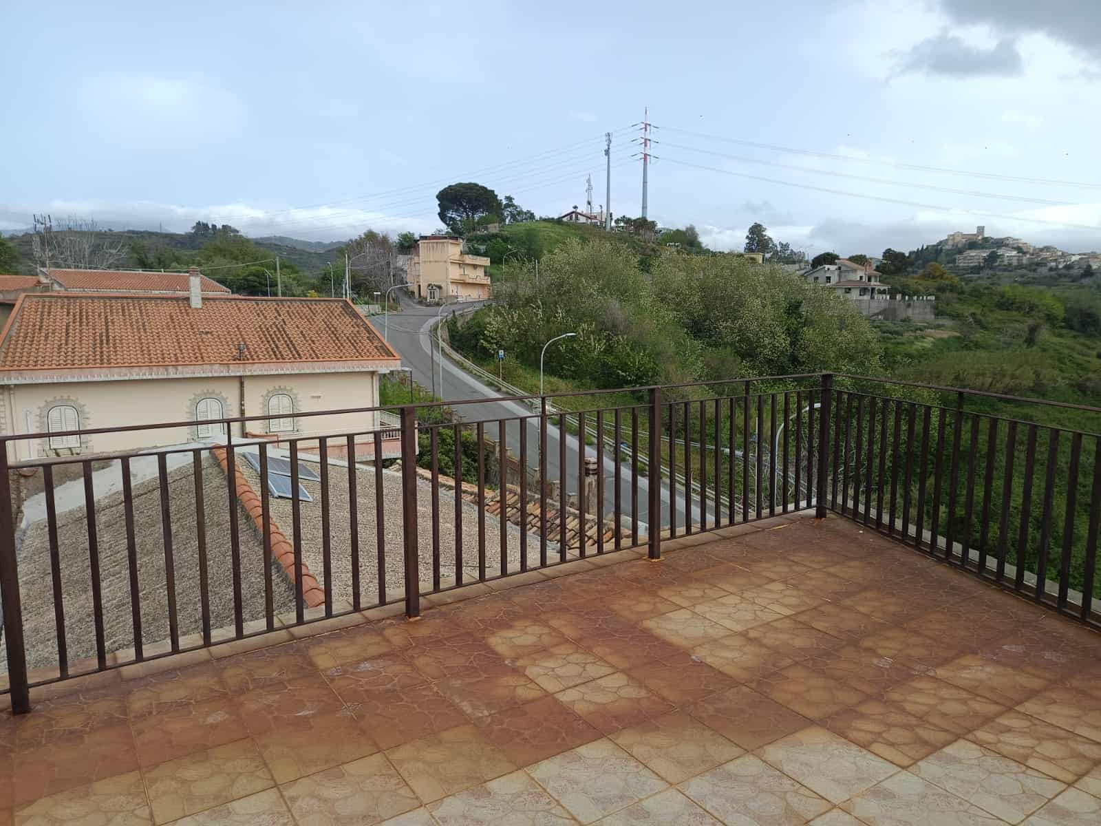 57f8655d-20e6-438a-805e-7e57a23f36d9 Vista panoramica dal balcone con ringhiera in metallo, case con tetti in terracotta e paesaggi collinari verdi, strada e abitazioni in lontananza, cielo nuvoloso, ideale per immagini di immobili e vista panoramica.