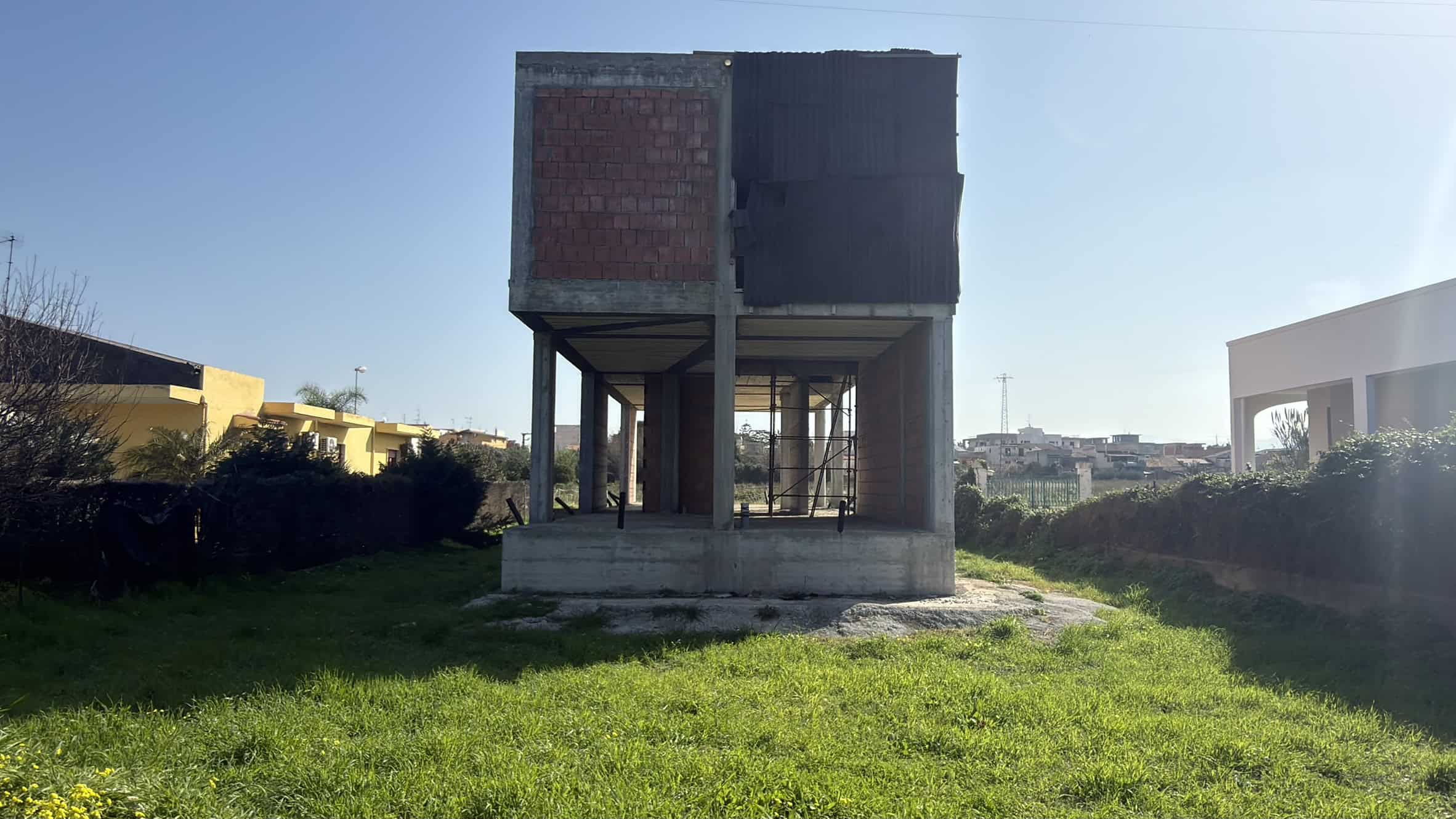 Pannello di costruzione di una casa in fase di realizzazione con struttura in cemento e mattoni, situata in un'area verde aperta sotto un cielo sereno.