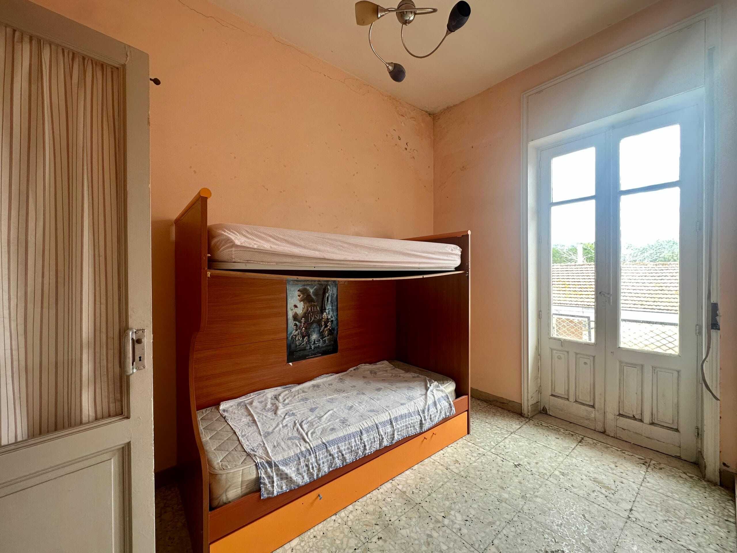 Letto a castello in camera con pareti rosa, finestra grande e porta finestra con vista su tetti e cielo, in stile semplice e funzionale, ideale per affitti o sistemazioni temporanee.