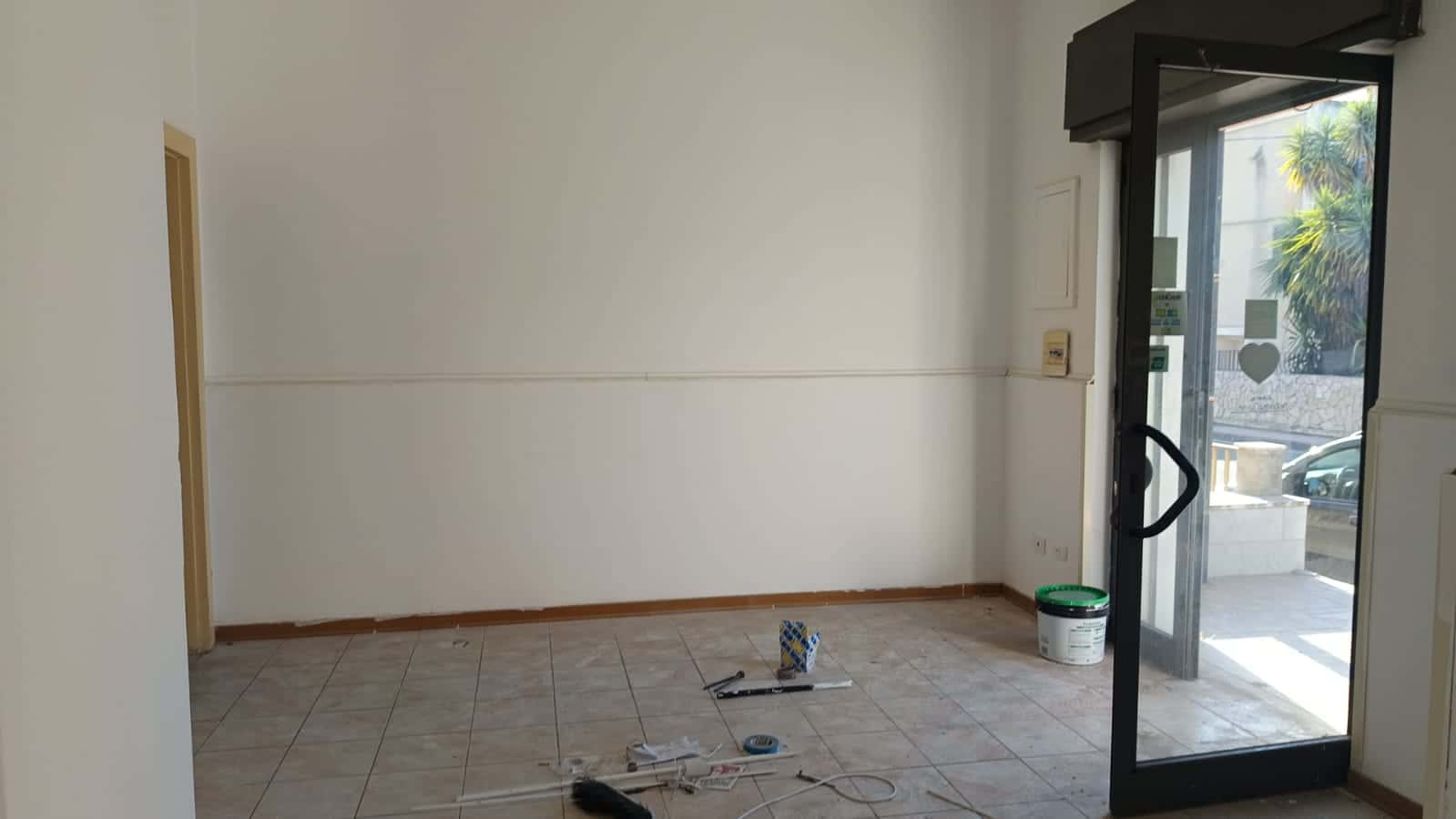 Immagine di un interno di negozio in fase di ristrutturazione con pareti bianche, pavimento in piastrelle e porta vetrata di accesso alla strada esterna.