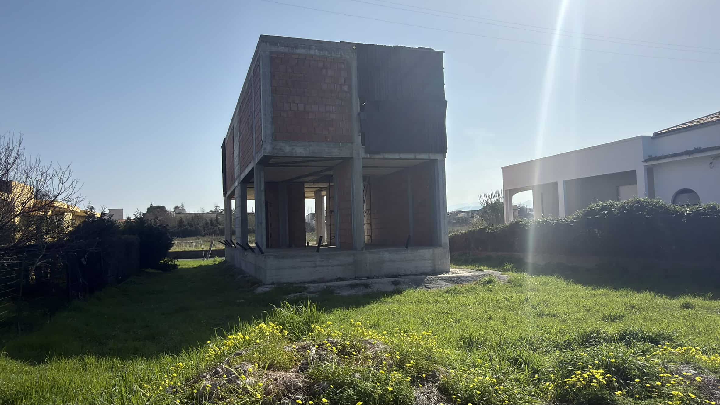 Cantiere edile con struttura in cemento e mattoni, pronto per la costruzione di una nuova abitazione o edificio commerciale. L'area circostante è verde e ben mantenuta, con un cielo sereno e sole bril.