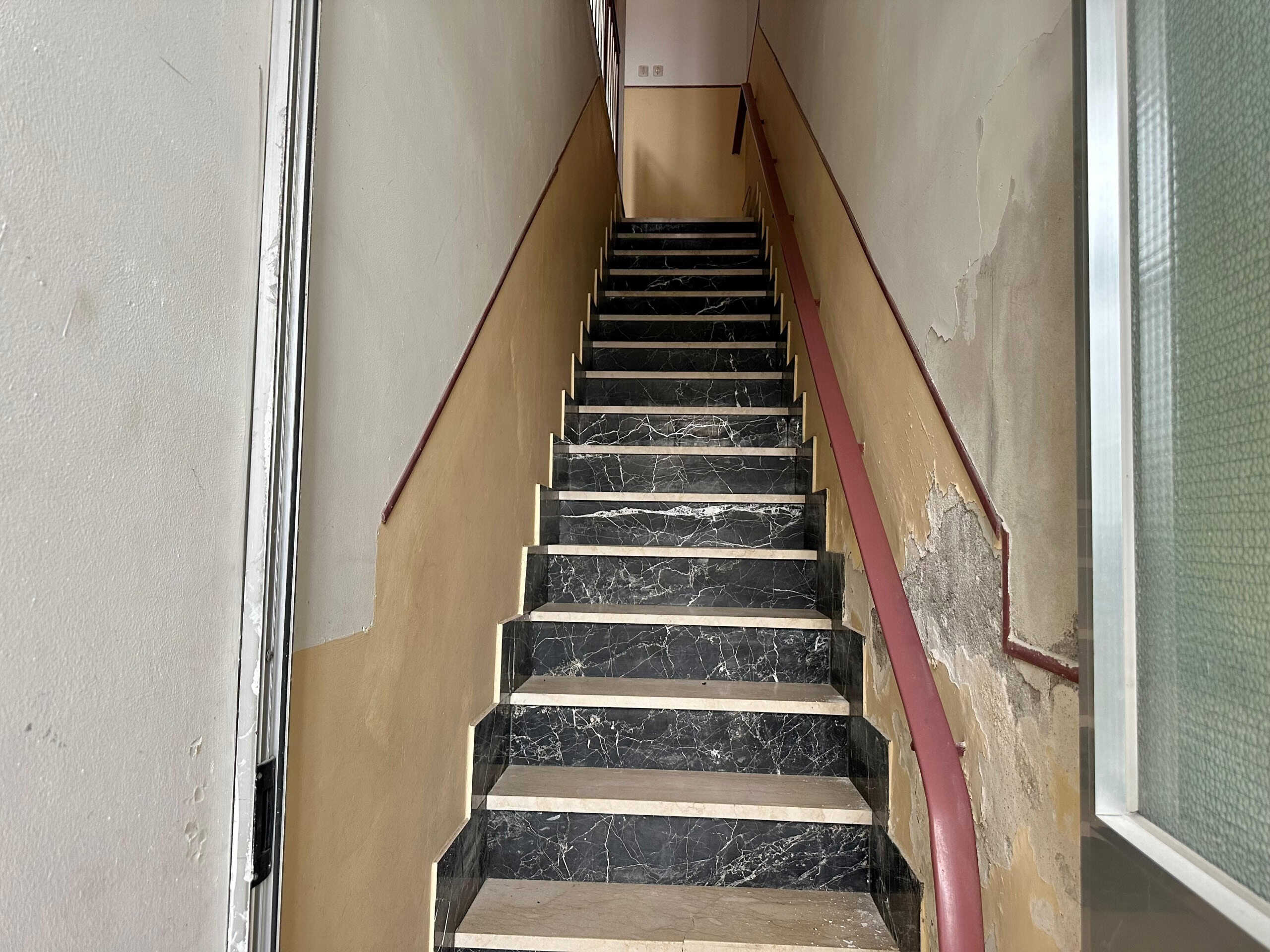 Scaletto in marmo nero e ringhiera in legno, all'interno di un edificio residenziale o commerciale, con pareti con pittura chipping e segni di deterioramento, creando un'immagine di ristrutturazione o manutenzione edilizia.