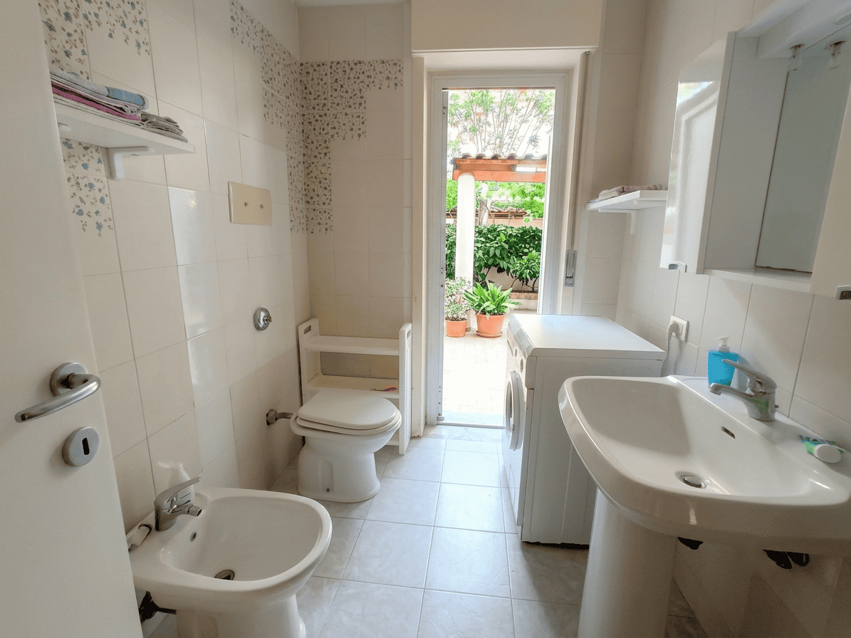 Bagno luminoso con accesso al giardino, dotato di lavabo, WC, bidet e lavatrice, in un appartamento in vendita in via Stazione 90, Oliveri, Me.