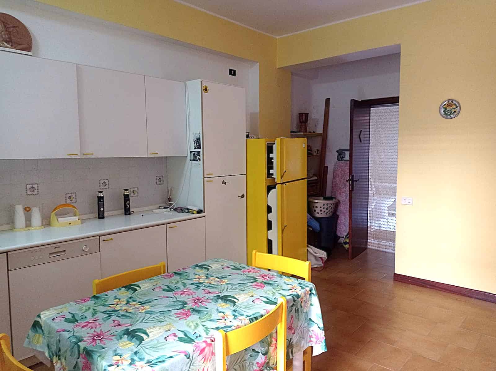 Lavastoviglie e cucina in stile classico con mobile bianco e dettagli in giallo, tavolo da pranzo con tovaglia floreale, ambiente luminoso con pareti gialle, zona giorno accogliente e funzionale.