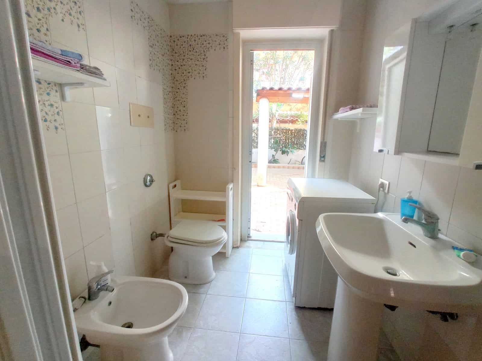 Bagno di casa con lavabo, WC e area lavanderia, luce naturale e accesso a spazio esterno, rivestimenti in piastrelle chiara, funzionale e luminosa.
