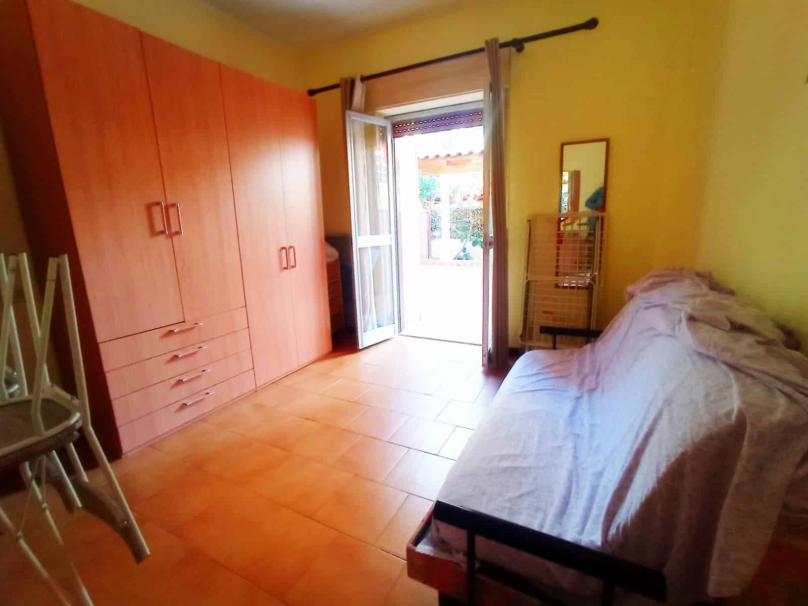 Armadio a muro in legno chiaro, soggiorno con divano coperto da lenzuola, porta finestra che apre su un balcone, pareti gialle, ambiente luminoso e accogliente, arredamento semplice e funzionale per case in affitto o vendita.
