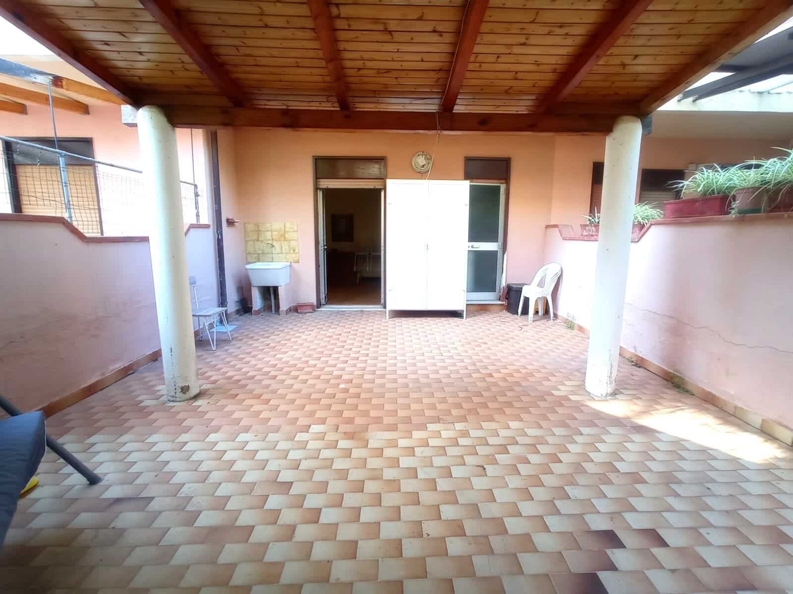 Spazio esterno con pavimentazione in mattonelle, tettoia in legno e pareti color pesca, con sedie e piante in vaso, ideale per relax e socializzazione in casa.
