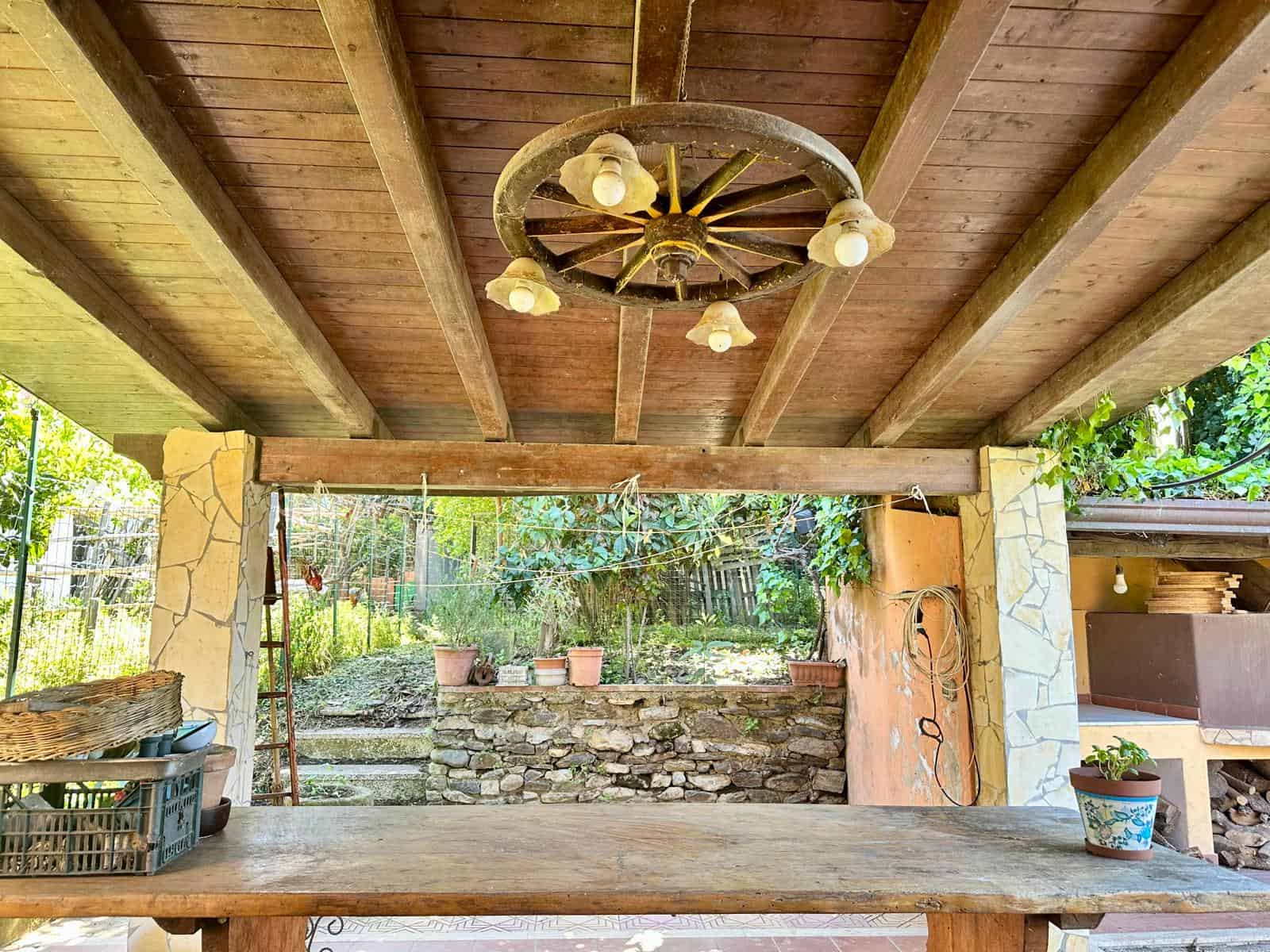 Antico lampadario a impianto wagon con luci a sfera, soffitto in legno e illuminazione calda, zona coperta all'aperto con tavolo in legno, piante in vaso e vista sul giardino rustico.