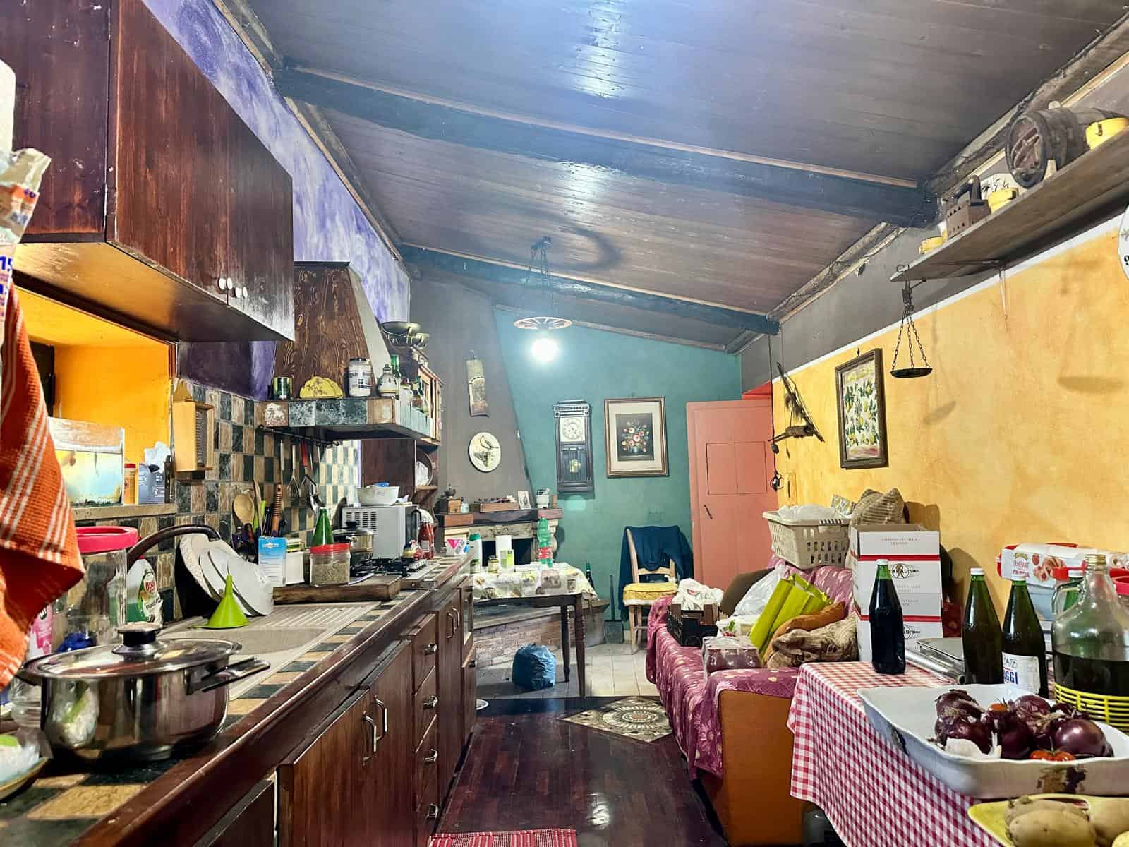 Casalinga cucina con soffitto in legno colorato, mobili in legno scuro, pareti verde e gialla, tavolo con tovaglia a scacchi rossi, vari oggetti e cibo, ambiente accogliente e informale.
